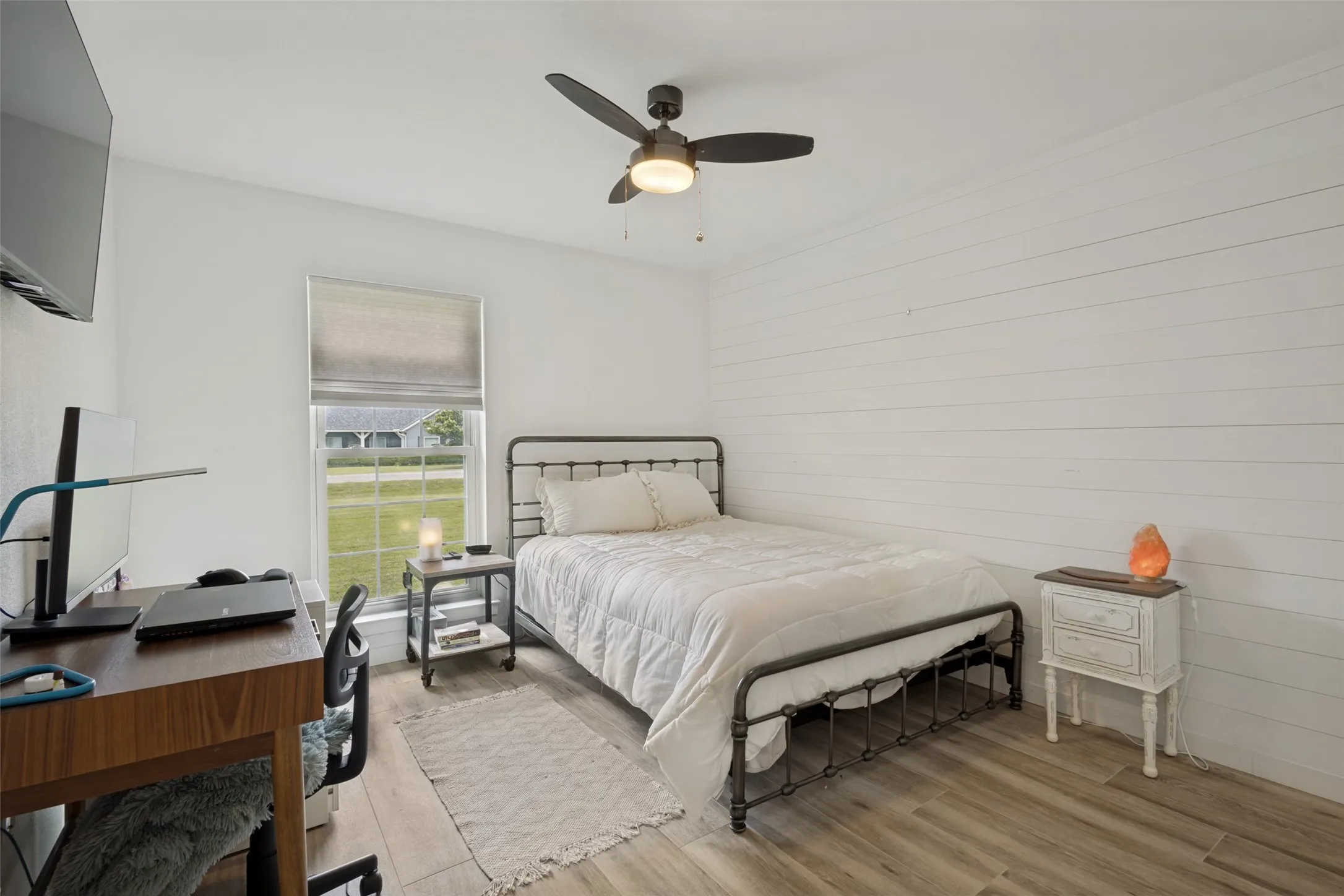White shiplap accent wall contrasting with smooth white walls, wood-finish flooring, and a ceiling fan with integrated lighting