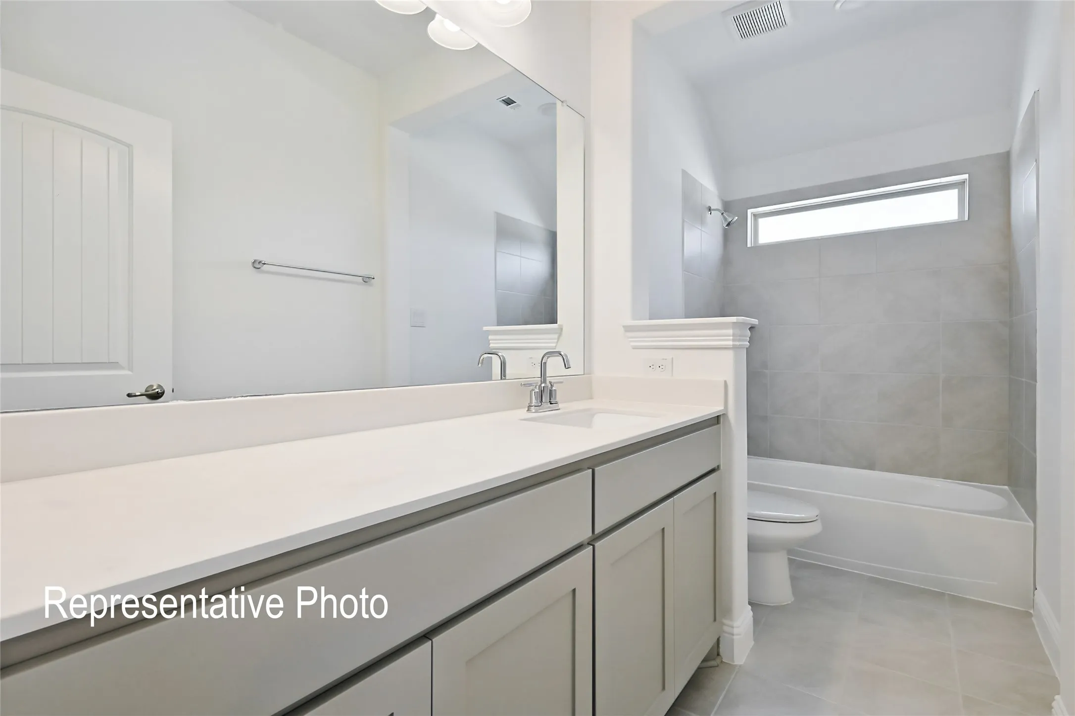 Bathroom featuring a light gray vanity with white countertop, chrome fixtures, a full-width mirror, and a tub-shower combination with gray tile surround and a horizontal window