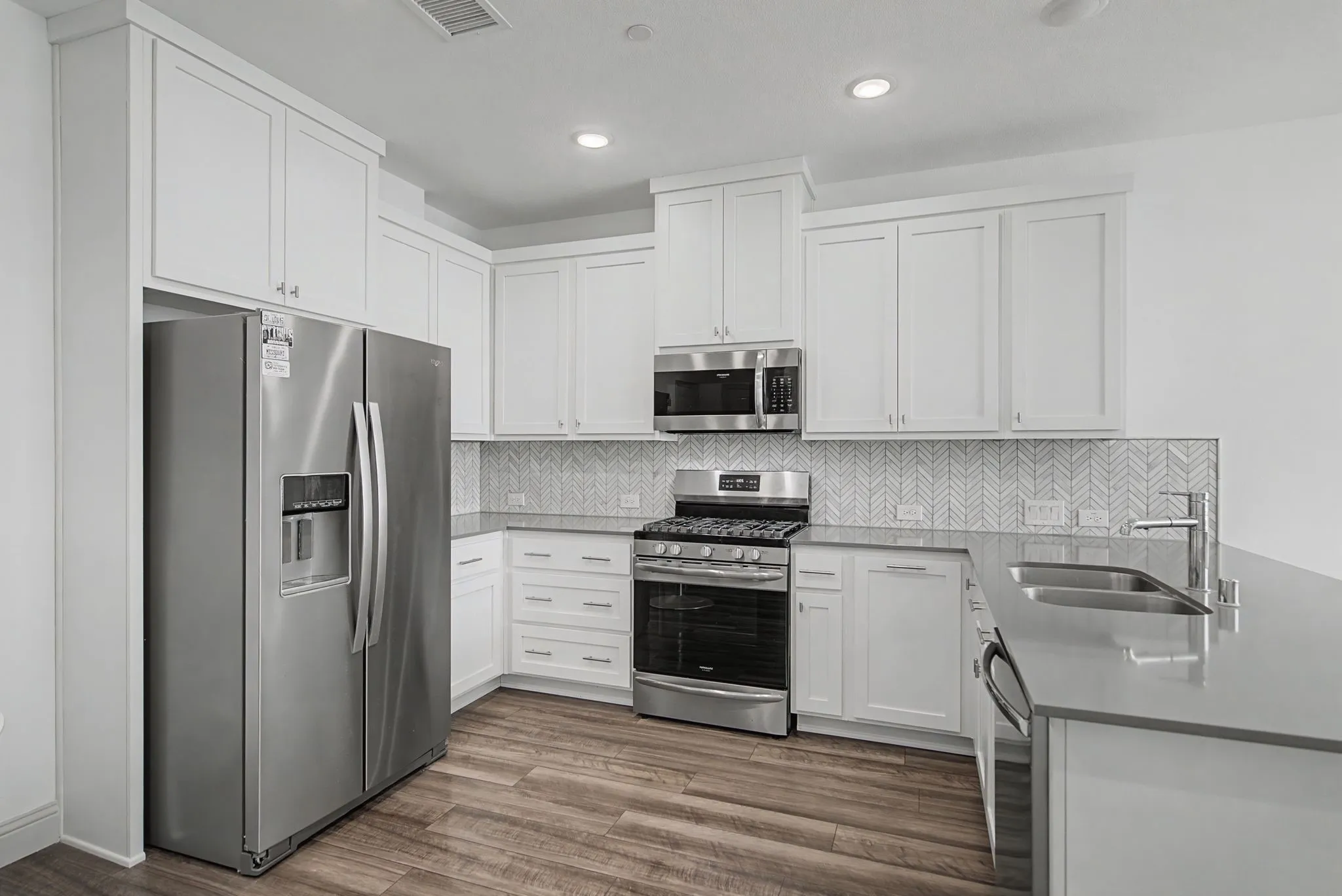 Kitchen featuring stainless steel appliances, white shaker cabinetry, wood-finish flooring, light-toned countertops, and a herringbone tile backsplash