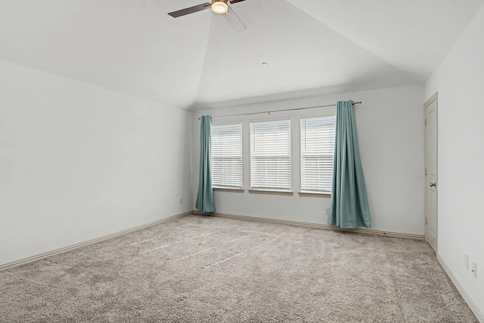 Spacious room featuring vaulted ceilings, plush carpeting, three windows with blinds, and a ceiling fan with integrated lighting