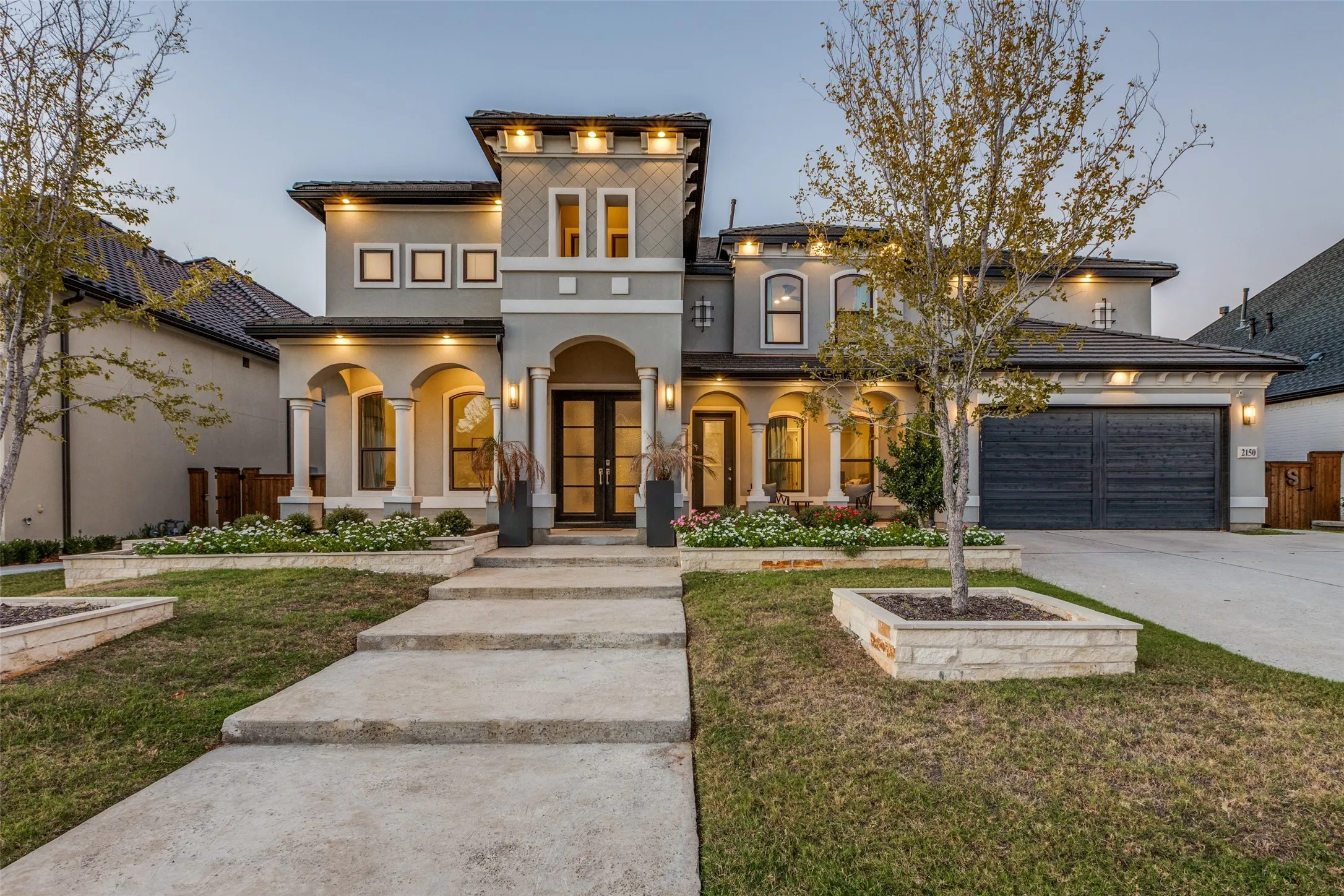 Contemporary stucco and stone exterior featuring a covered entry porch with arches, a double-door entry, a two-car garage, and tiered landscaping