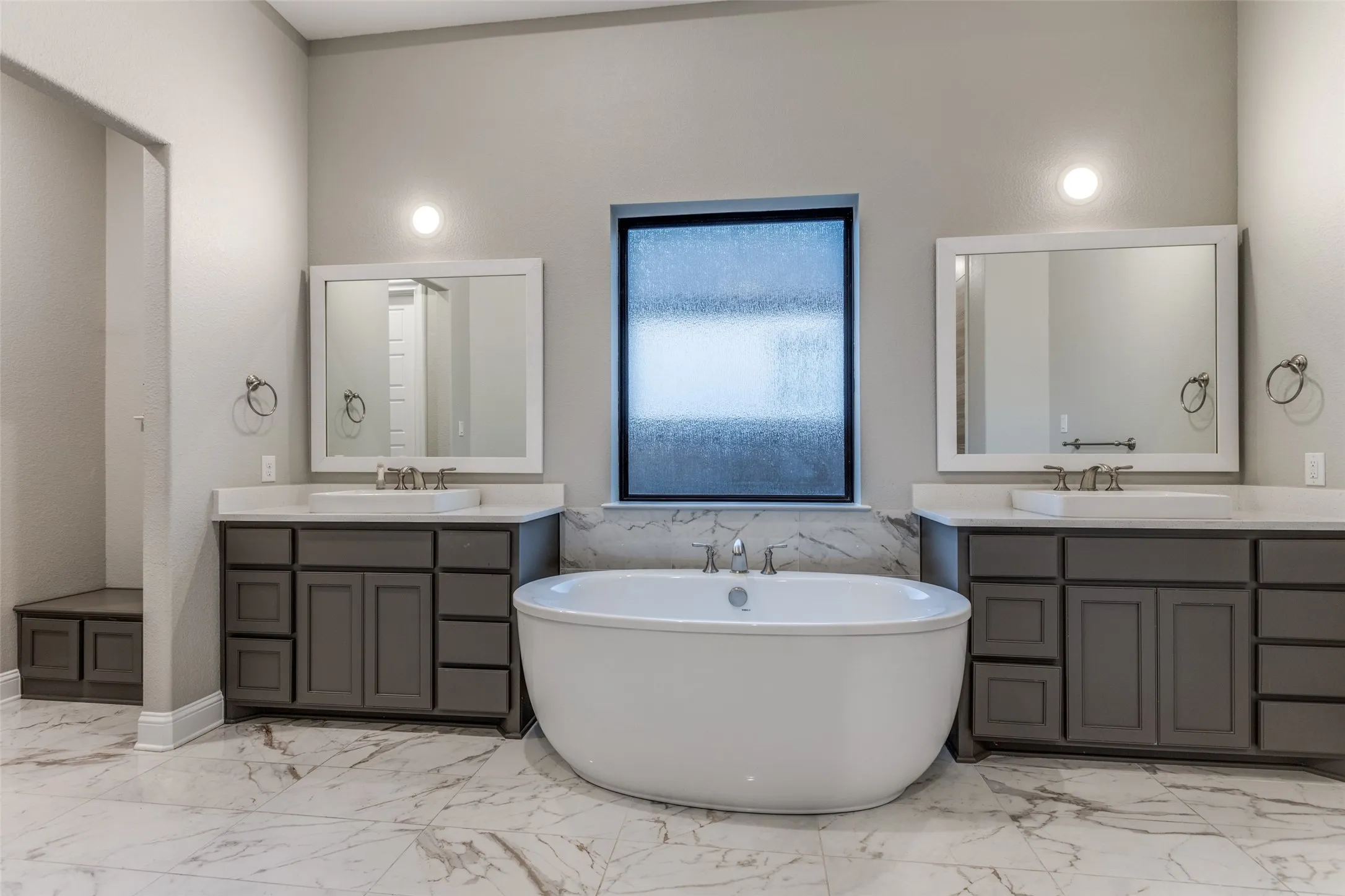 Freestanding soaking tub centered between dual vanities with grey cabinetry and white countertops