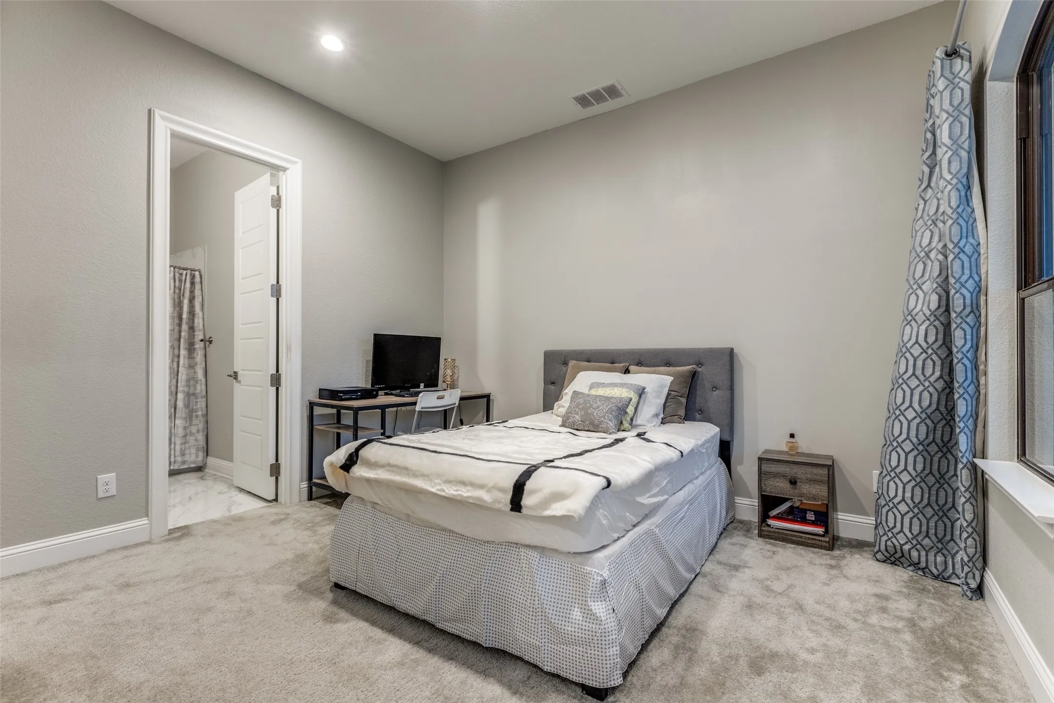 Carpeted room with light gray walls, an open doorway revealing a tiled bathroom, and a window with patterned drapes