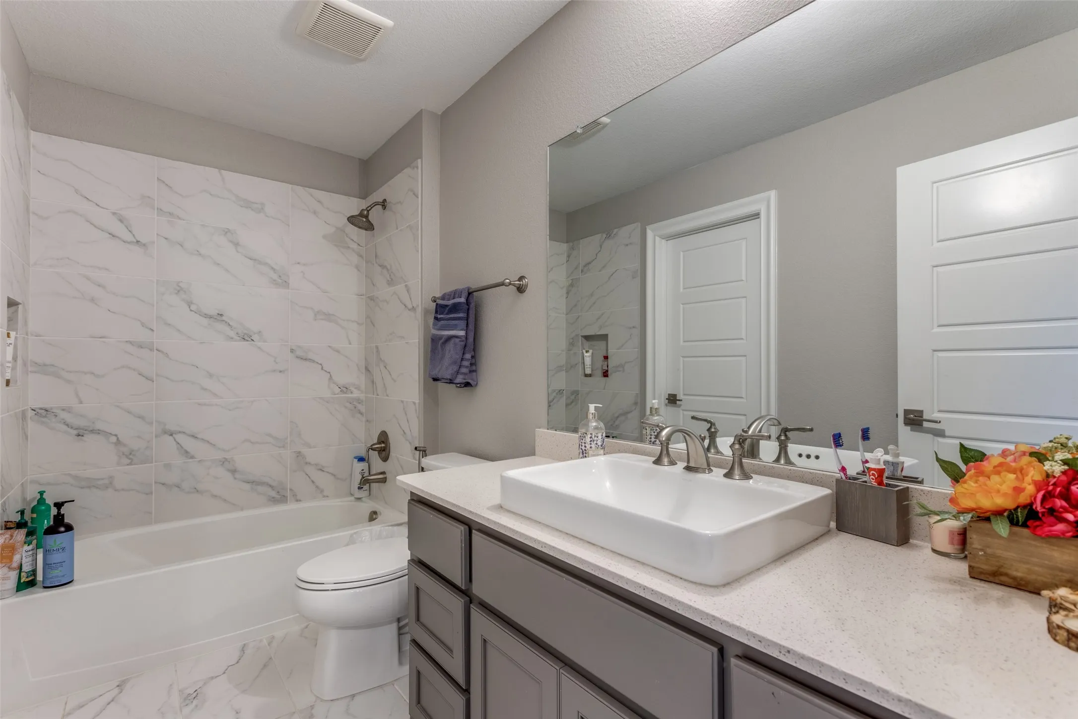 Bathroom featuring a white rectangular vessel sink, gray vanity cabinetry, light speckled countertop, and a white tiled shower surround with a built-in niche
