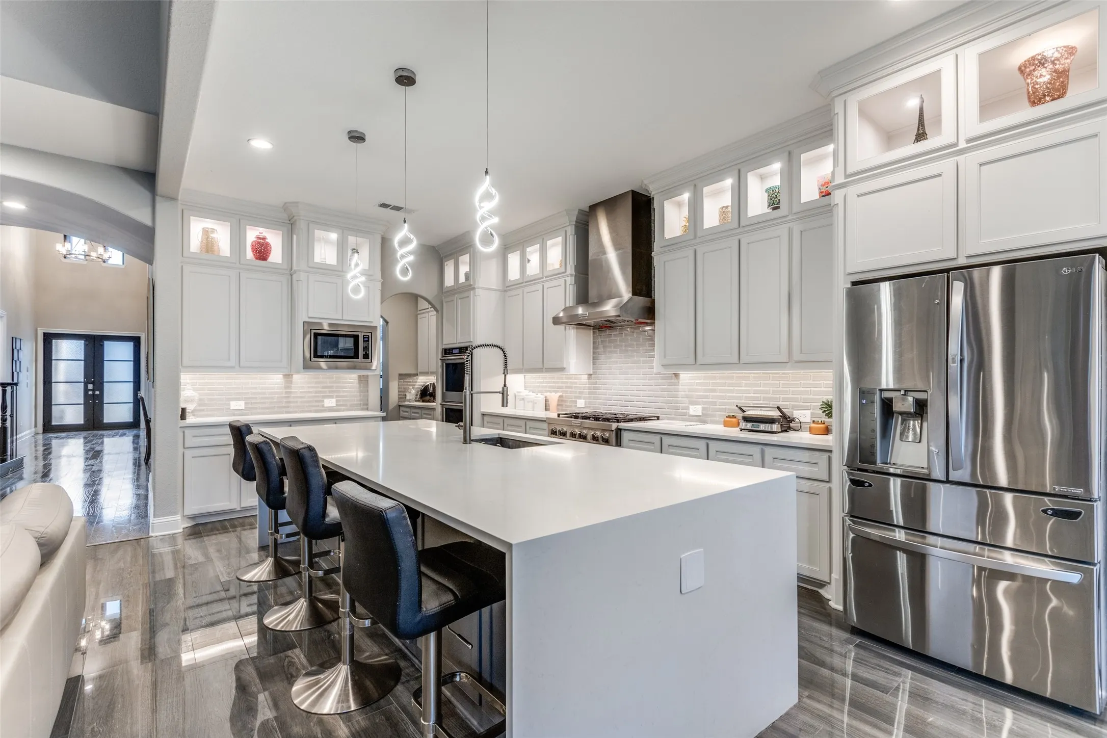 Spacious kitchen featuring a large central island, light-toned cabinetry with display shelves, stainless steel appliances, a tile backsplash, and dark wood-finish flooring