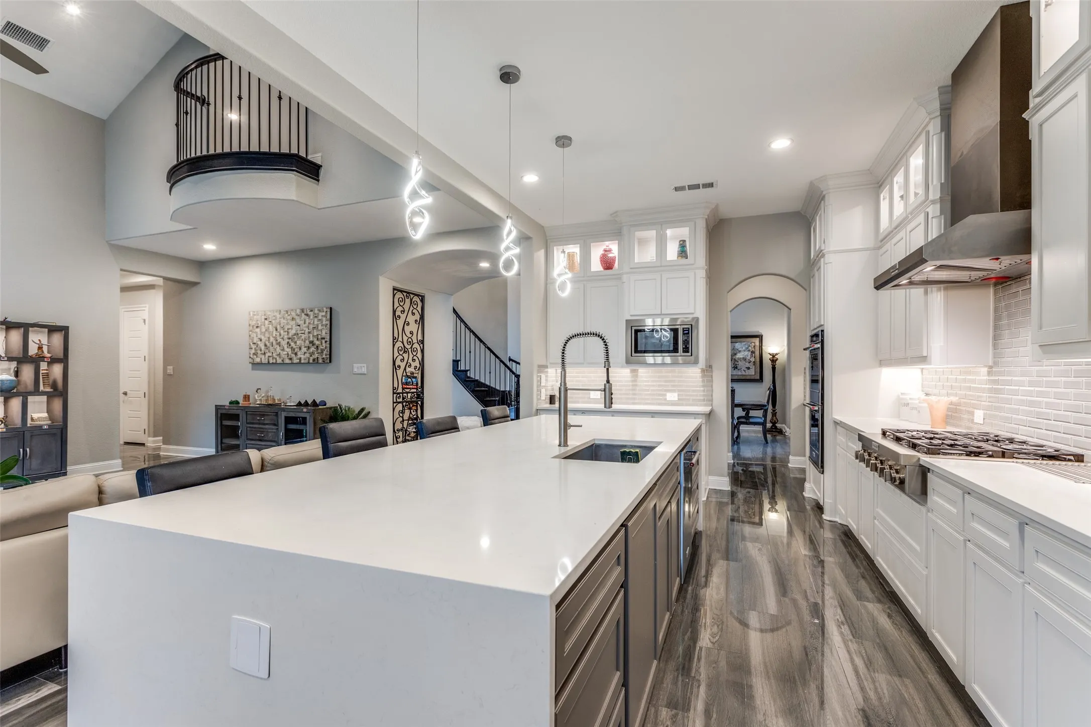 Open-concept kitchen featuring a large island with an undermount sink, built-in gas range with stainless steel hood, white cabinetry with display uppers, and wood-finish flooring throughout