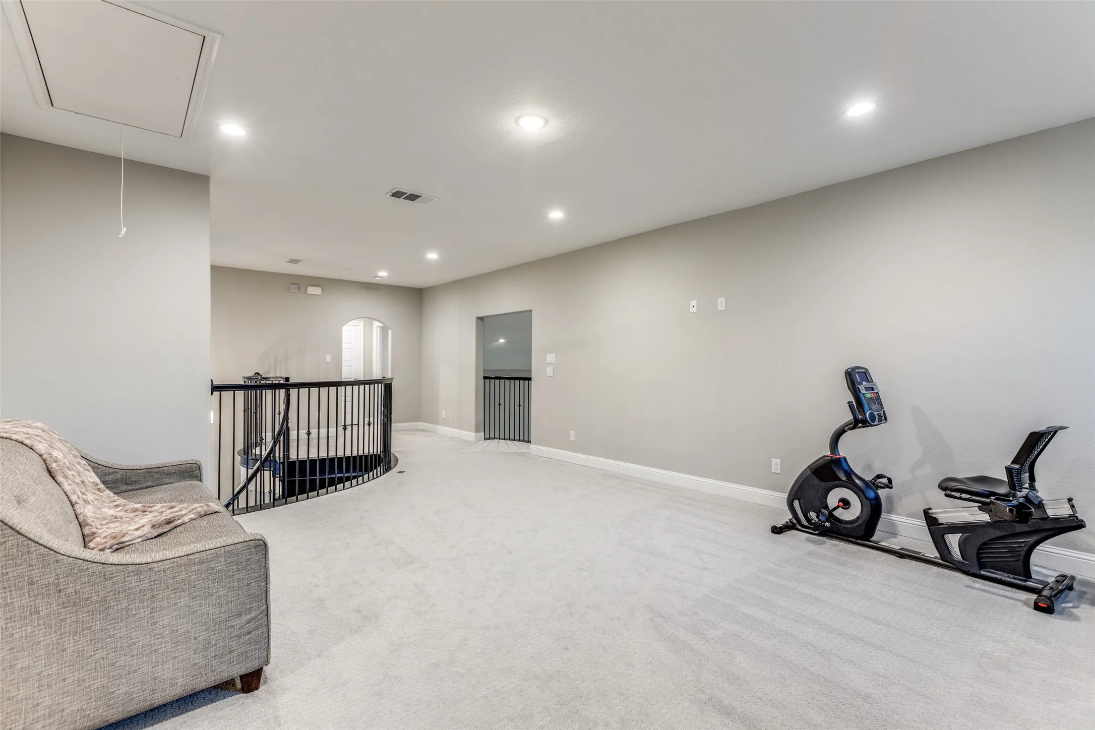 Spacious bonus room featuring light grey carpeting, recessed lighting, and a wrought iron banister overlooking a curved staircase