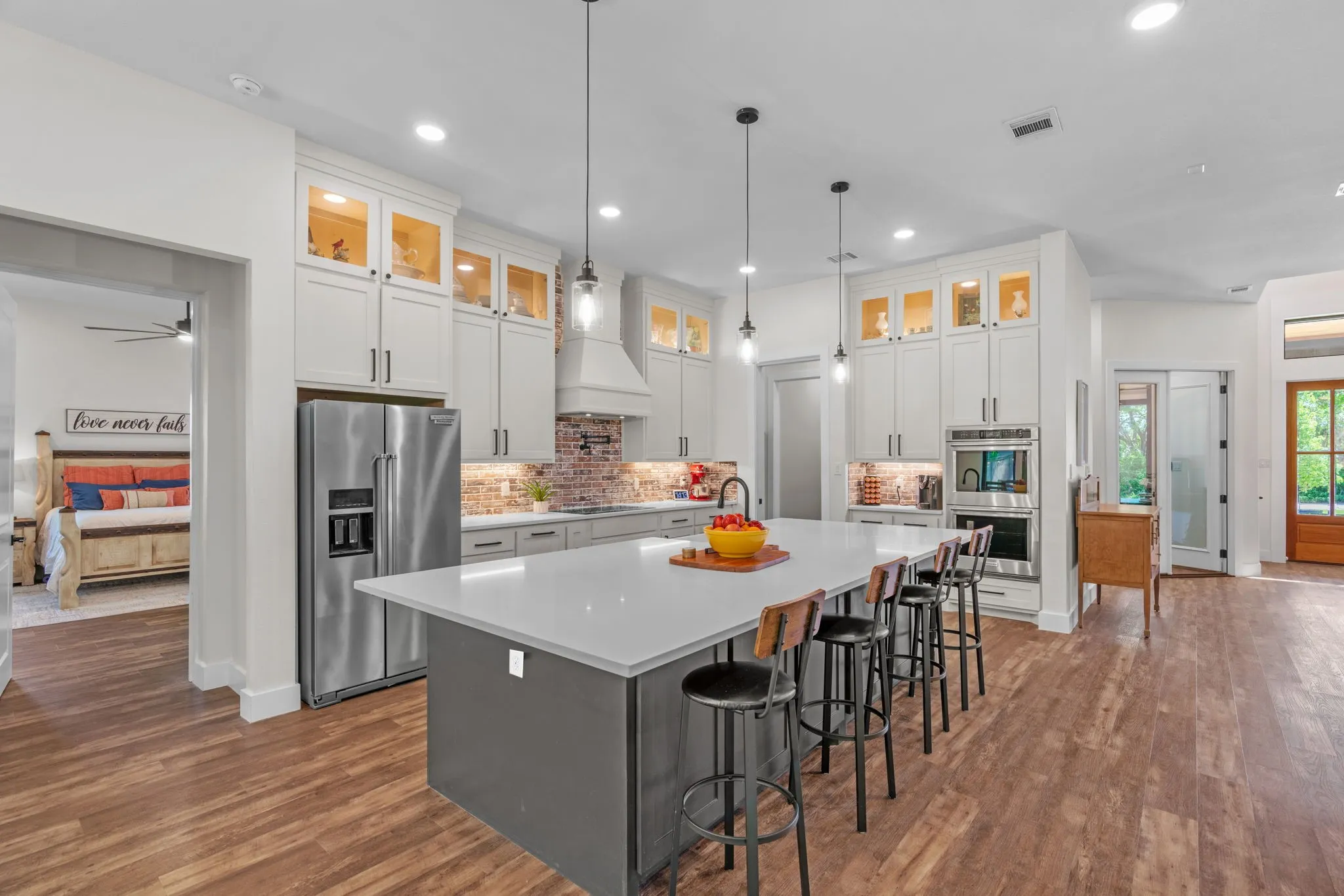 Gourmet kitchen featuring a large central island with seating, stainless steel appliances, white cabinetry with upper glass display cabinets, brick-finish backsplash, and wood-finish flooring