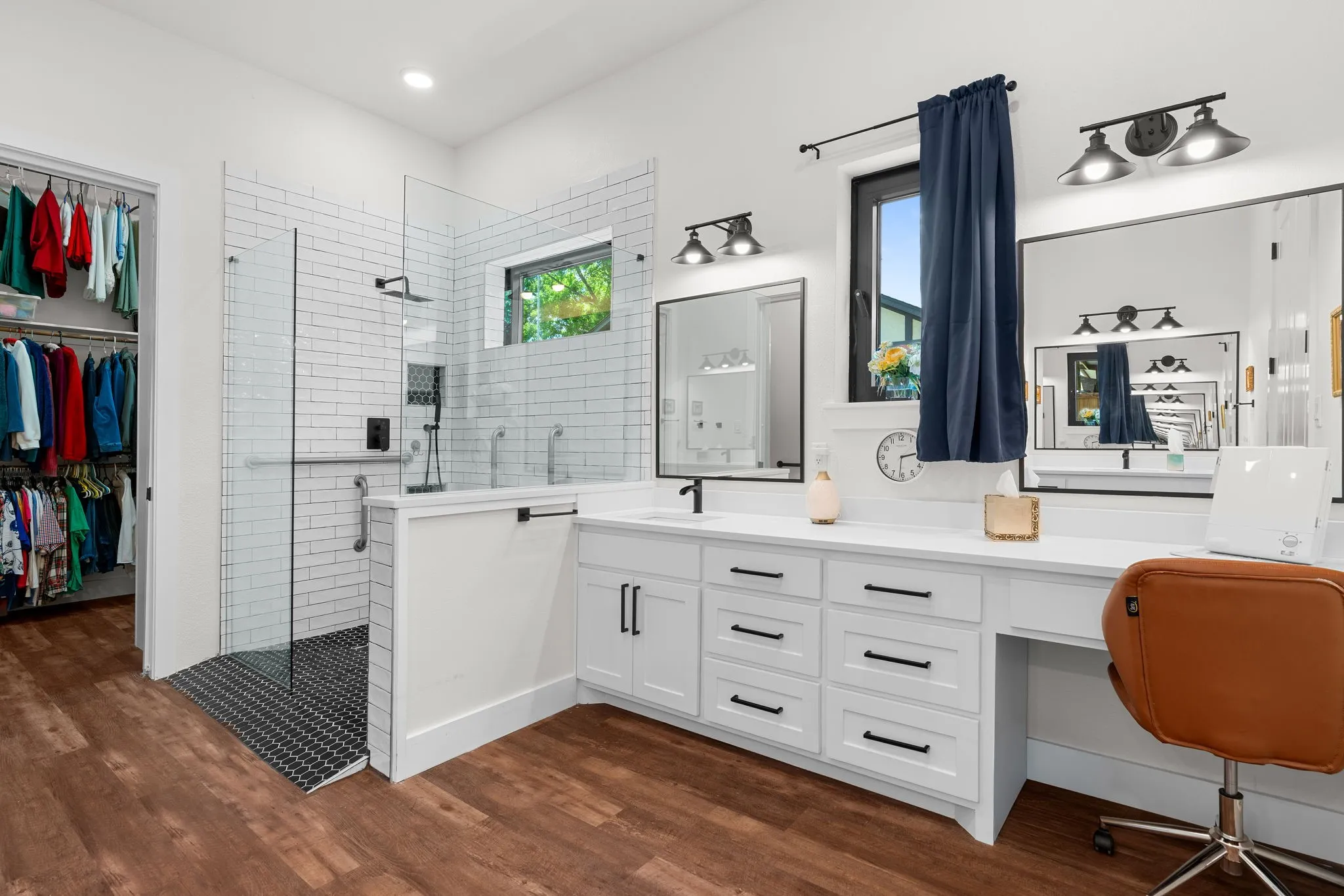 Spacious bathroom featuring a glass-enclosed shower with white subway tile, an expansive double vanity with white cabinetry and dark hardware, wood-finish flooring, and a walk-in closet