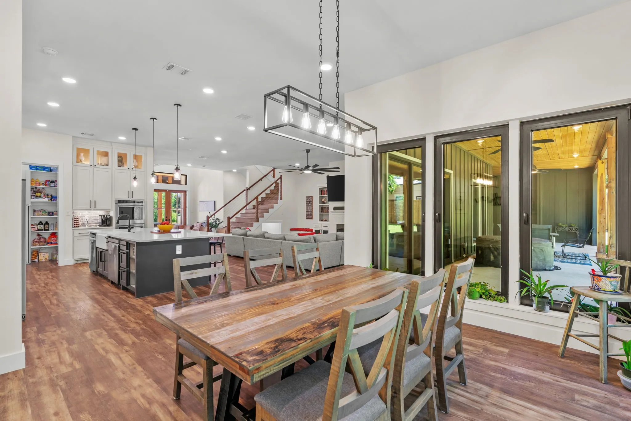 Open-concept interior featuring wood-finish flooring throughout, a modern kitchen with a large island, a dining area with a linear chandelier, and sliding glass doors opening to an outdoor patio