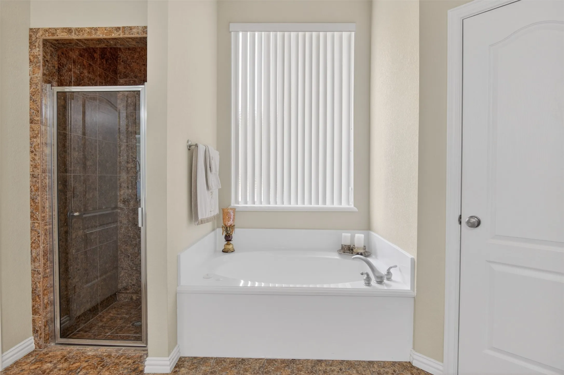 Soaking tub with a window featuring vertical blinds, complemented by a separate shower enclosure with a glass door and stone-finish tile surround