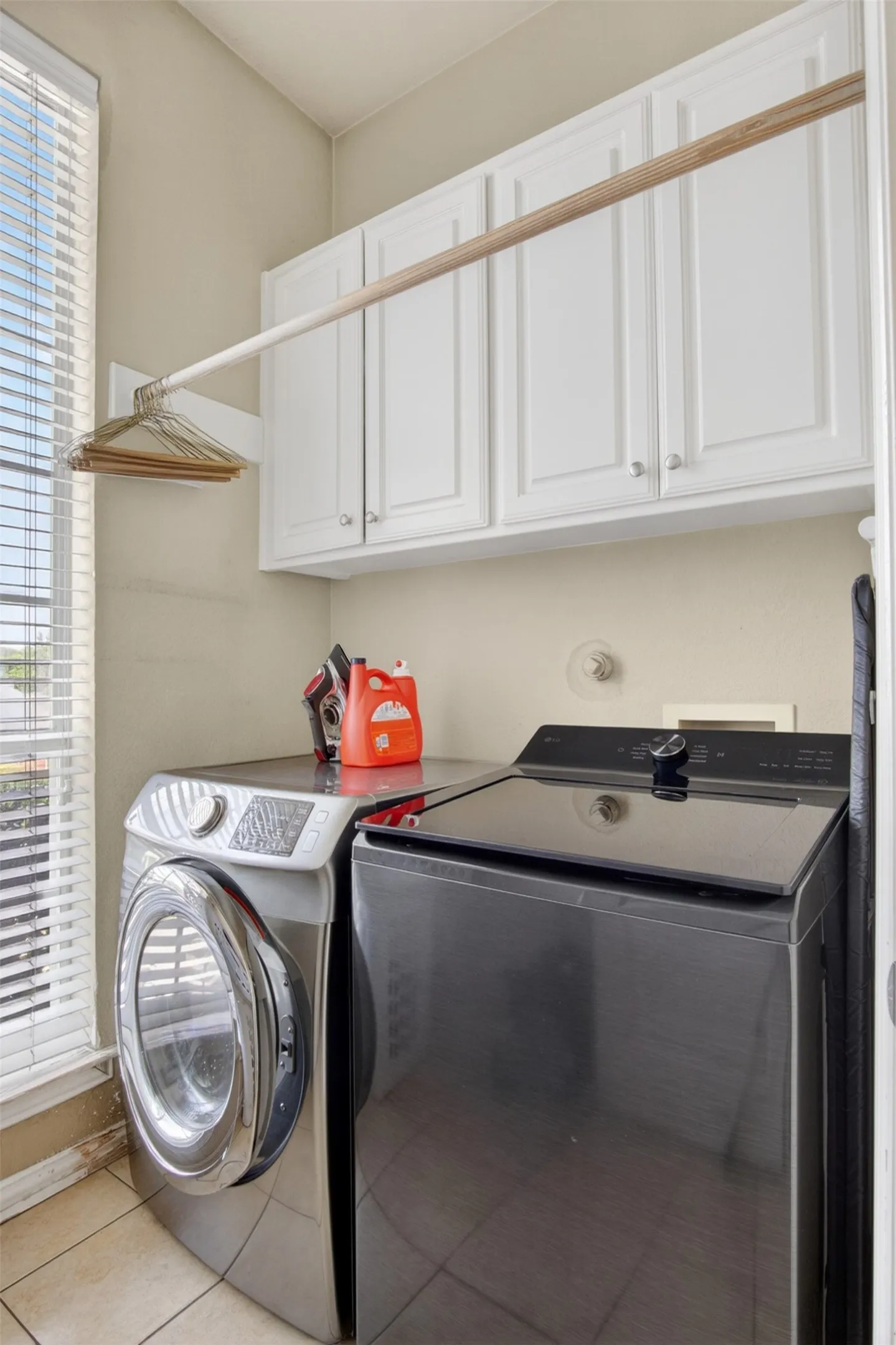 Dedicated laundry area featuring ceramic tile flooring, upper cabinetry with white shaker-style doors, a built-in hanging rod, and natural light from the window
