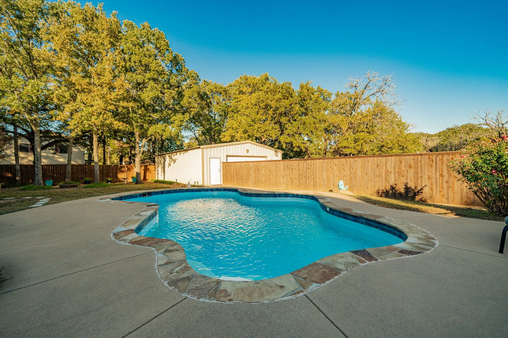 Freeform swimming pool with stone coping, surrounded by a concrete deck and a wood privacy fence