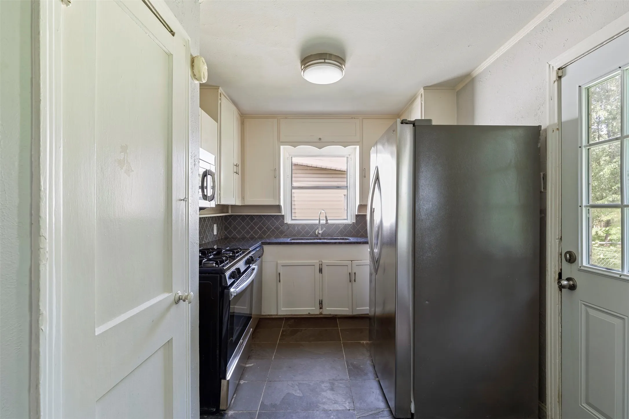 Galley kitchen featuring white cabinetry, dark countertops, diamond pattern backsplash, stainless steel appliances, and a glass-paned exterior door