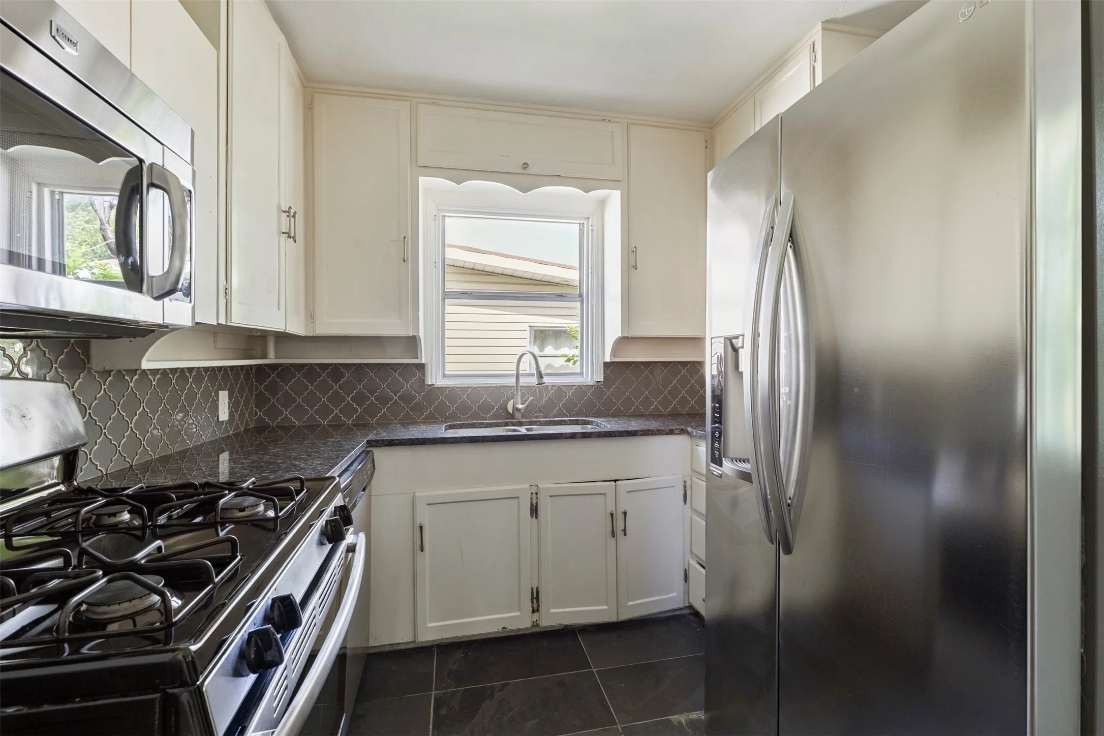 Compact kitchen featuring white cabinetry, dark countertops, dark tile flooring, a geometric tile backsplash, and stainless steel appliances