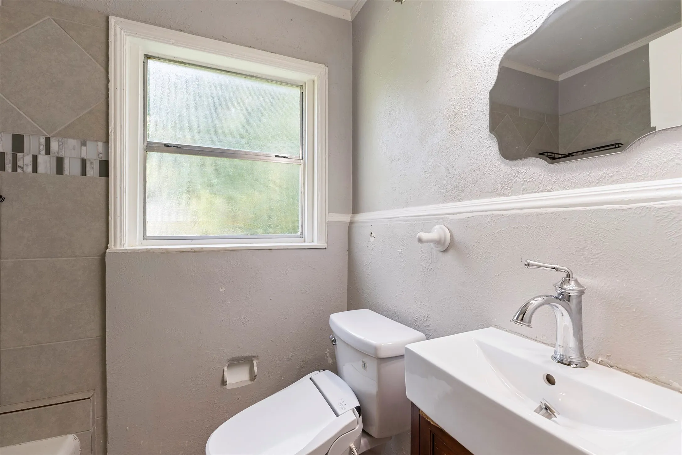 Bathroom featuring a single window with privacy glass, tiled shower surround with decorative accent band, floating vanity with vessel sink, wall-mounted mirror, and chair rail molding
