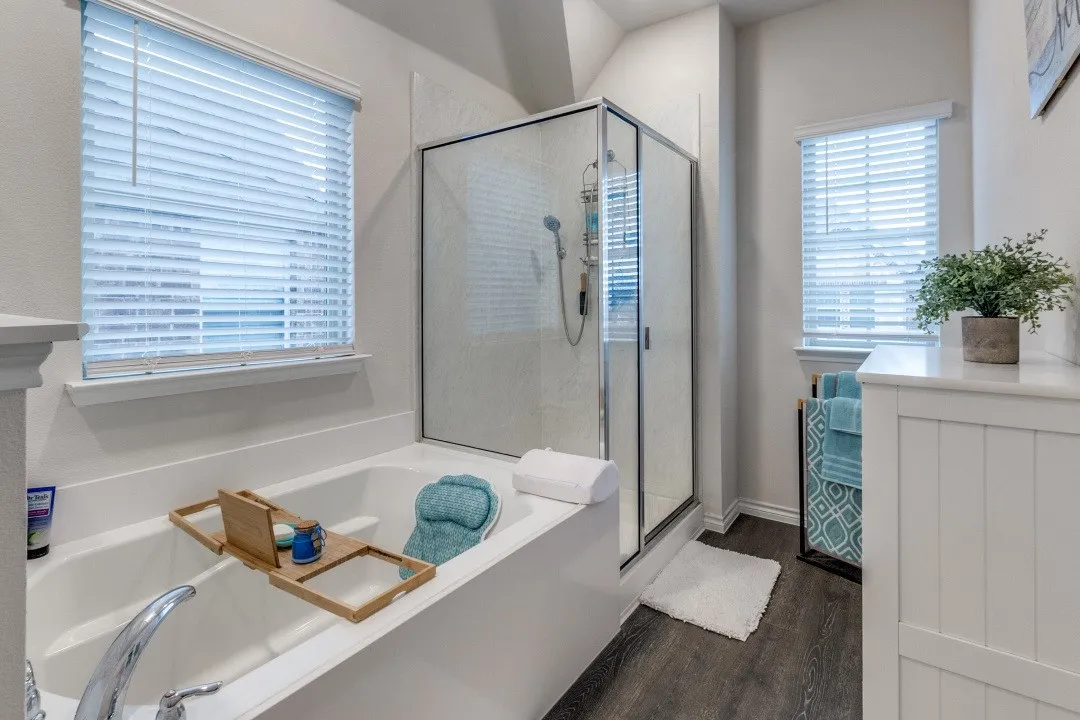 Soaking tub with a polished chrome faucet, glass-enclosed shower, white window blinds, wood-finish flooring, and a white paneled vanity