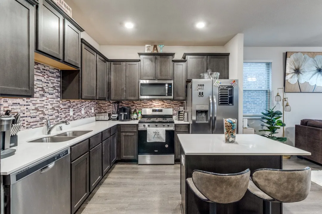 Modern kitchen featuring dark wood-finish cabinetry, stainless steel appliances, a mosaic tile backsplash, and a central island with a light-toned countertop