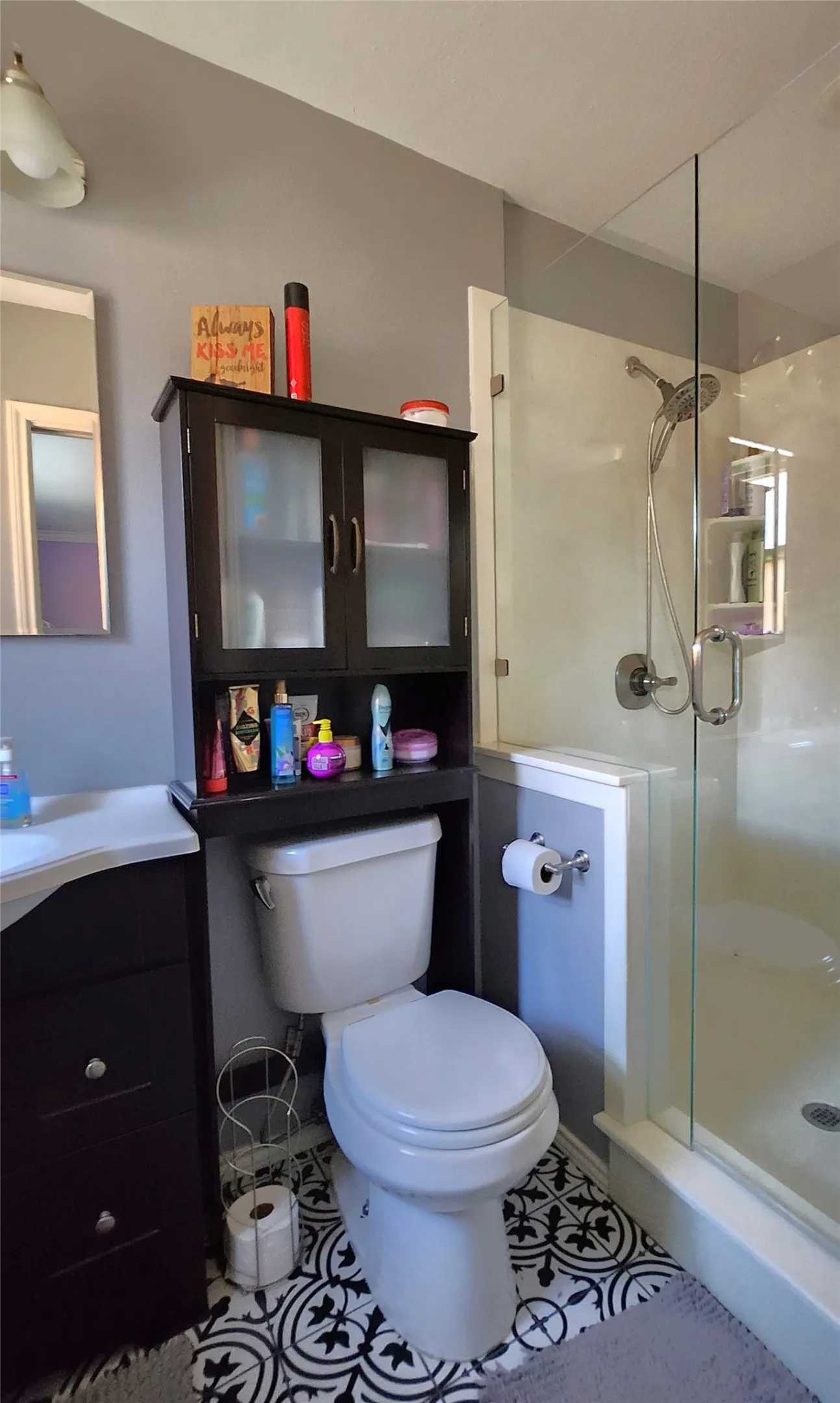 Bathroom featuring a glass-enclosed shower, patterned floor tile, over-the-toilet storage cabinetry, and a dark wood vanity with a white countertop