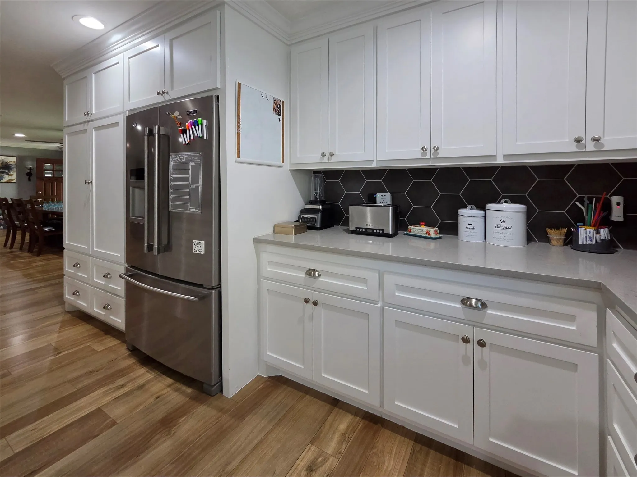 Well-appointed kitchen featuring white shaker cabinetry, stainless steel French door refrigerator, light-toned countertops, dark hexagonal tile backsplash, and wood-finish flooring