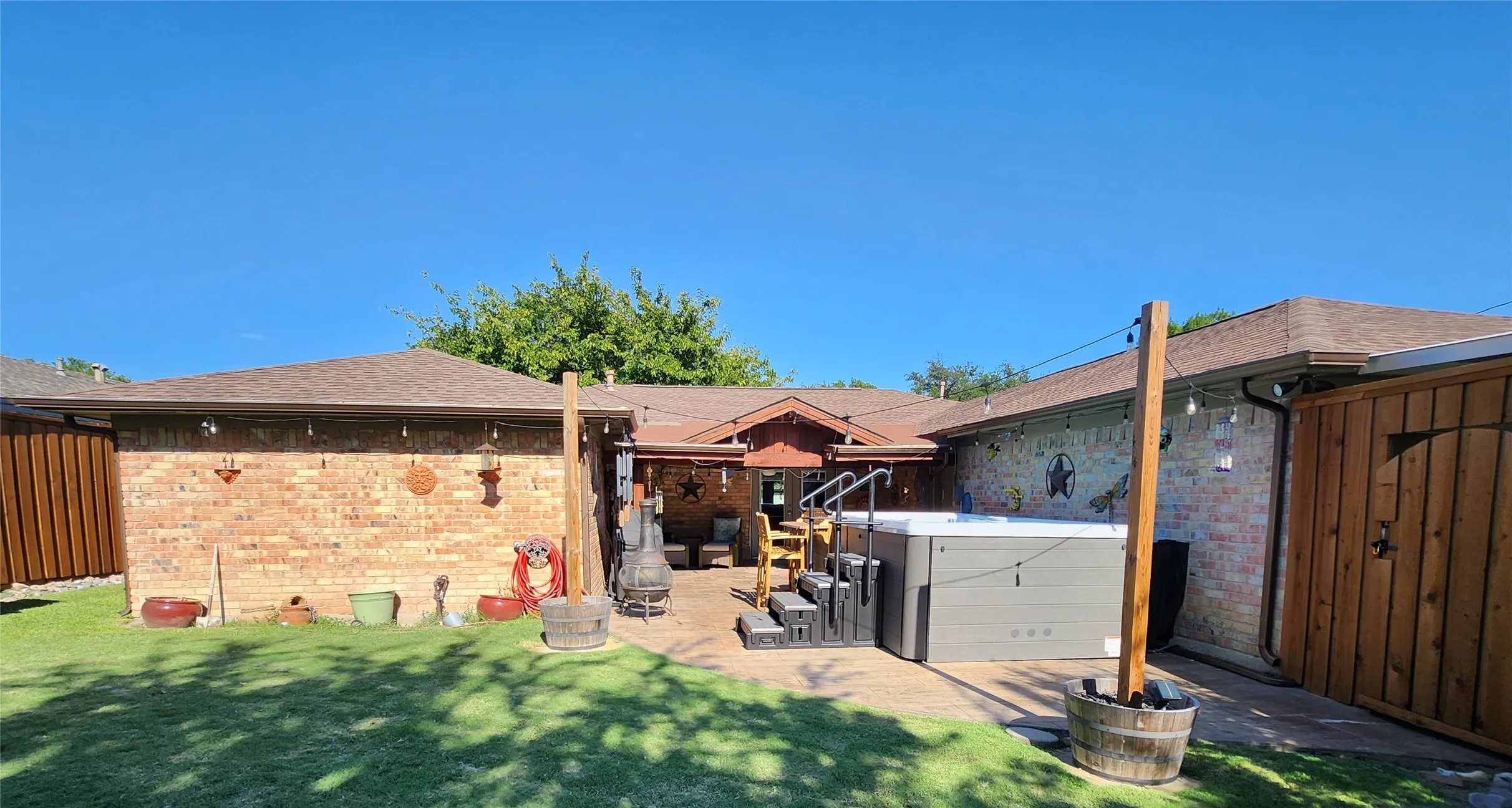 Private backyard featuring a hot tub with tiered steps, brick exterior walls, a covered patio area, and a wooden privacy fence
