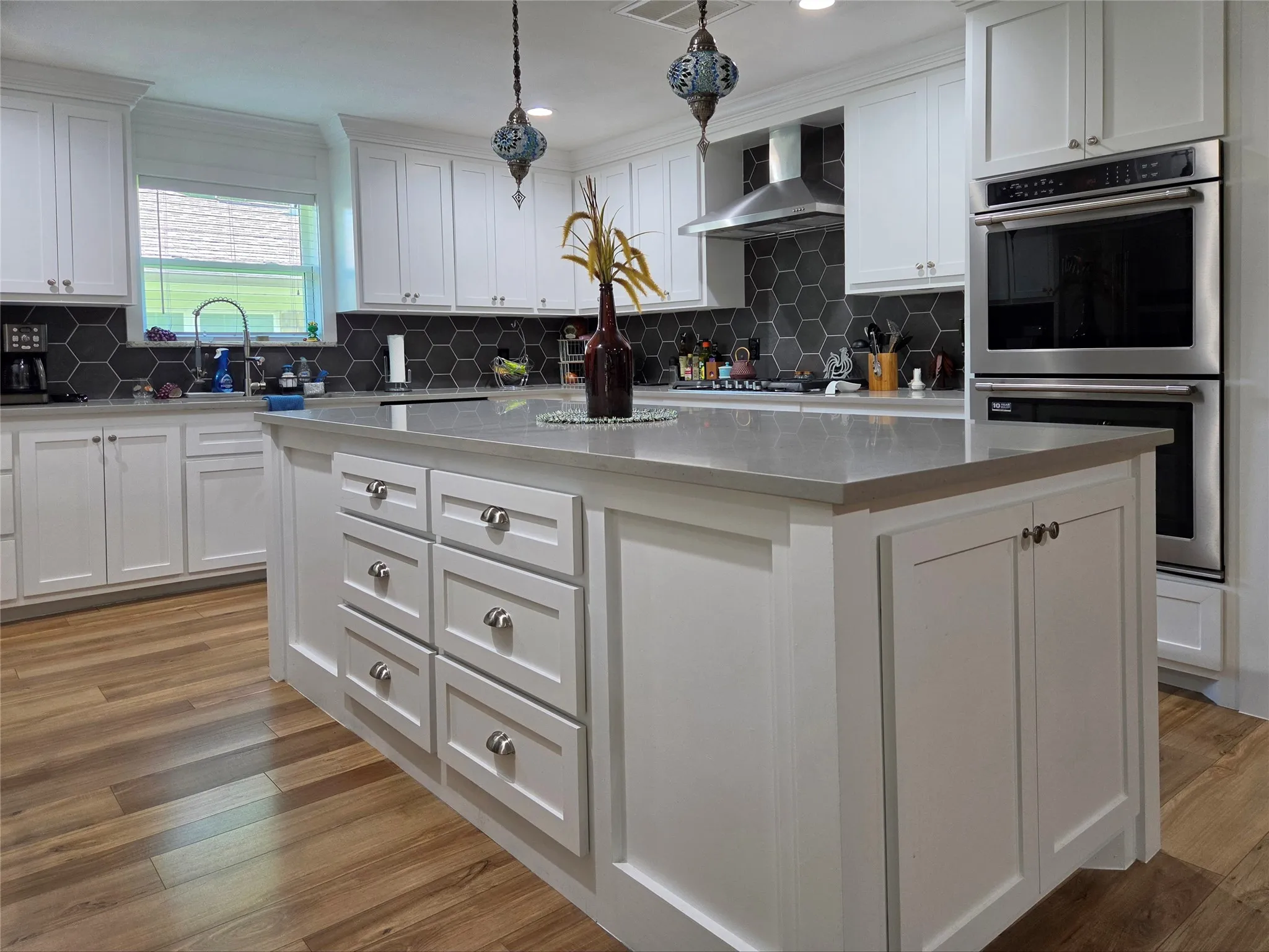 Kitchen island with a light-toned countertop, white cabinetry with silver hardware, wood-finish flooring, a dark hexagonal tile backsplash, and stainless steel double ovens