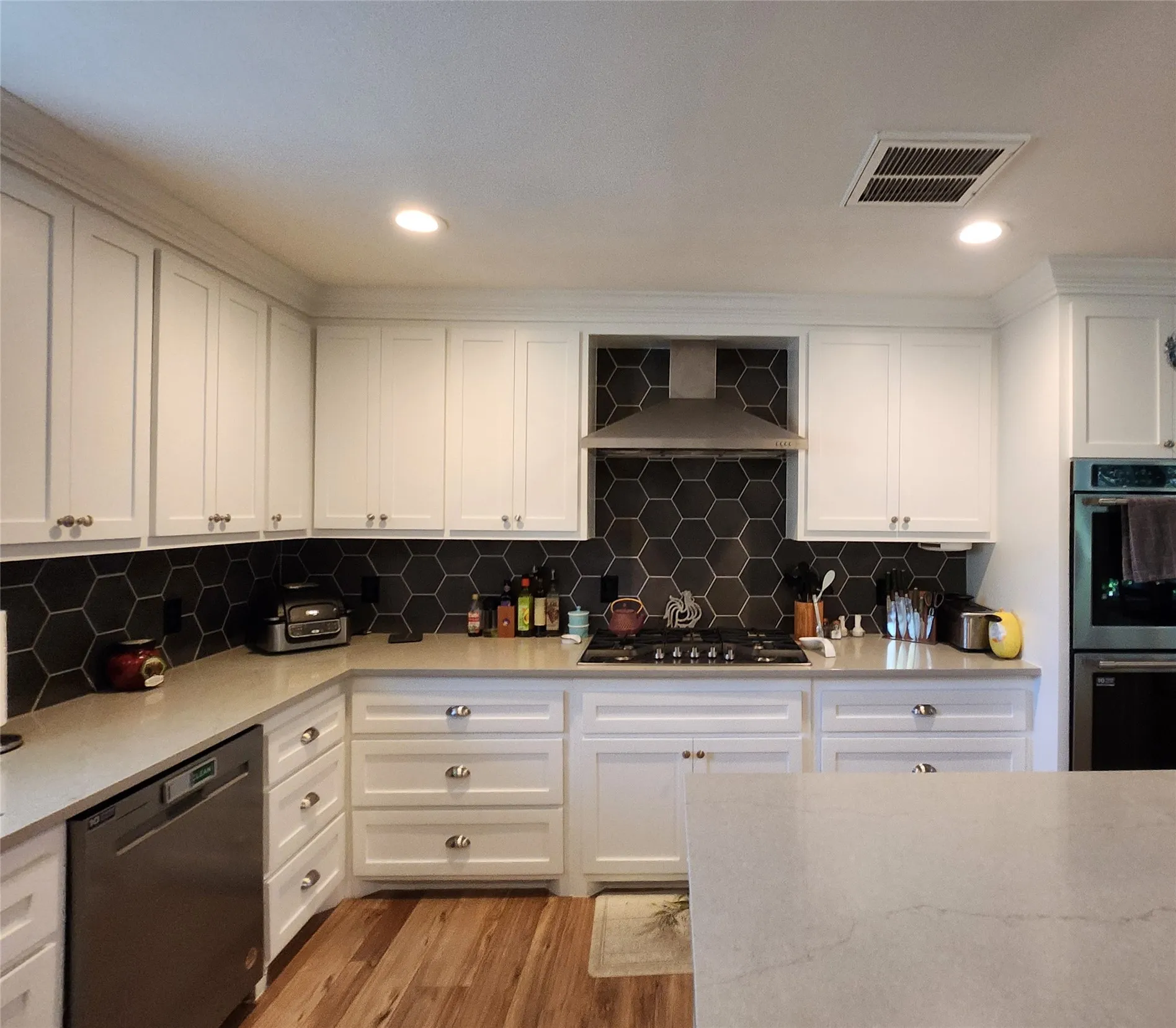 Kitchen featuring white shaker cabinetry, stainless steel appliances, dark hexagonal tile backsplash, light stone countertops, and wood-finish flooring