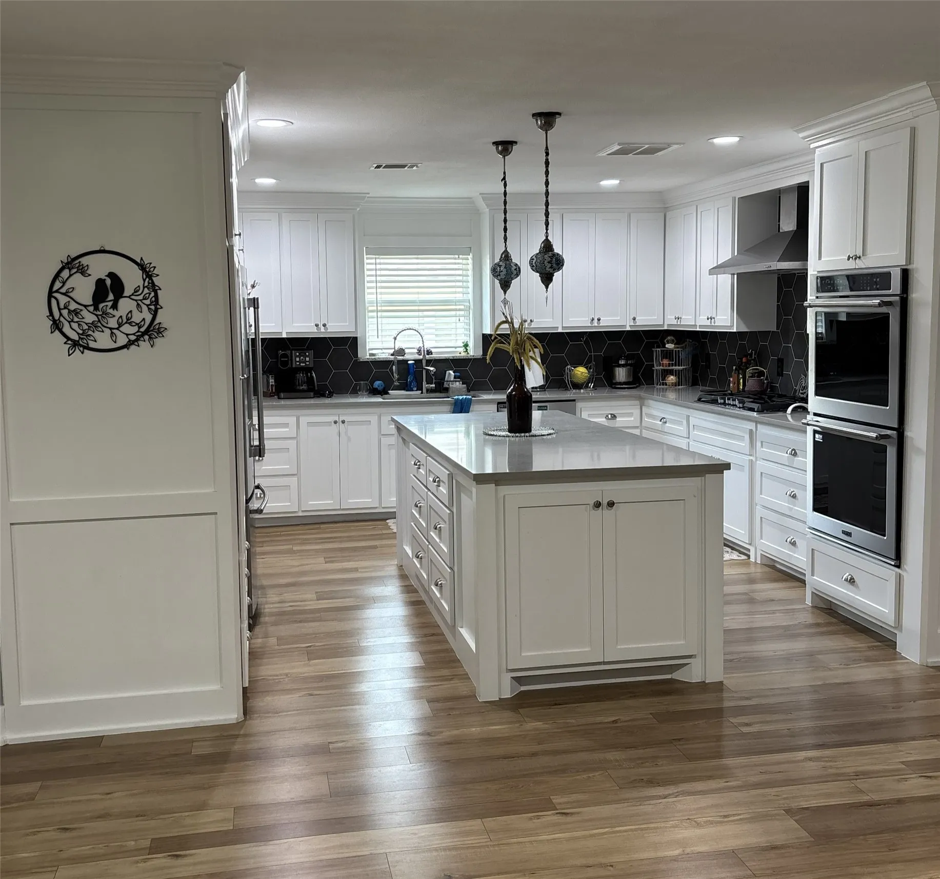 Spacious kitchen featuring white cabinetry, a large central island with a light-toned countertop, integrated stainless steel double ovens, and a dark hexagonal tile backsplash