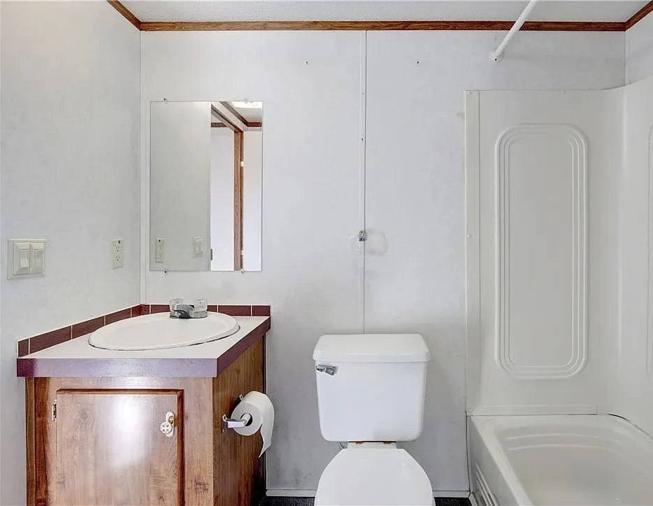 Bathroom featuring a wood-finish vanity with an oval sink, a wall-mounted mirror, a toilet, and a white shower/tub combination