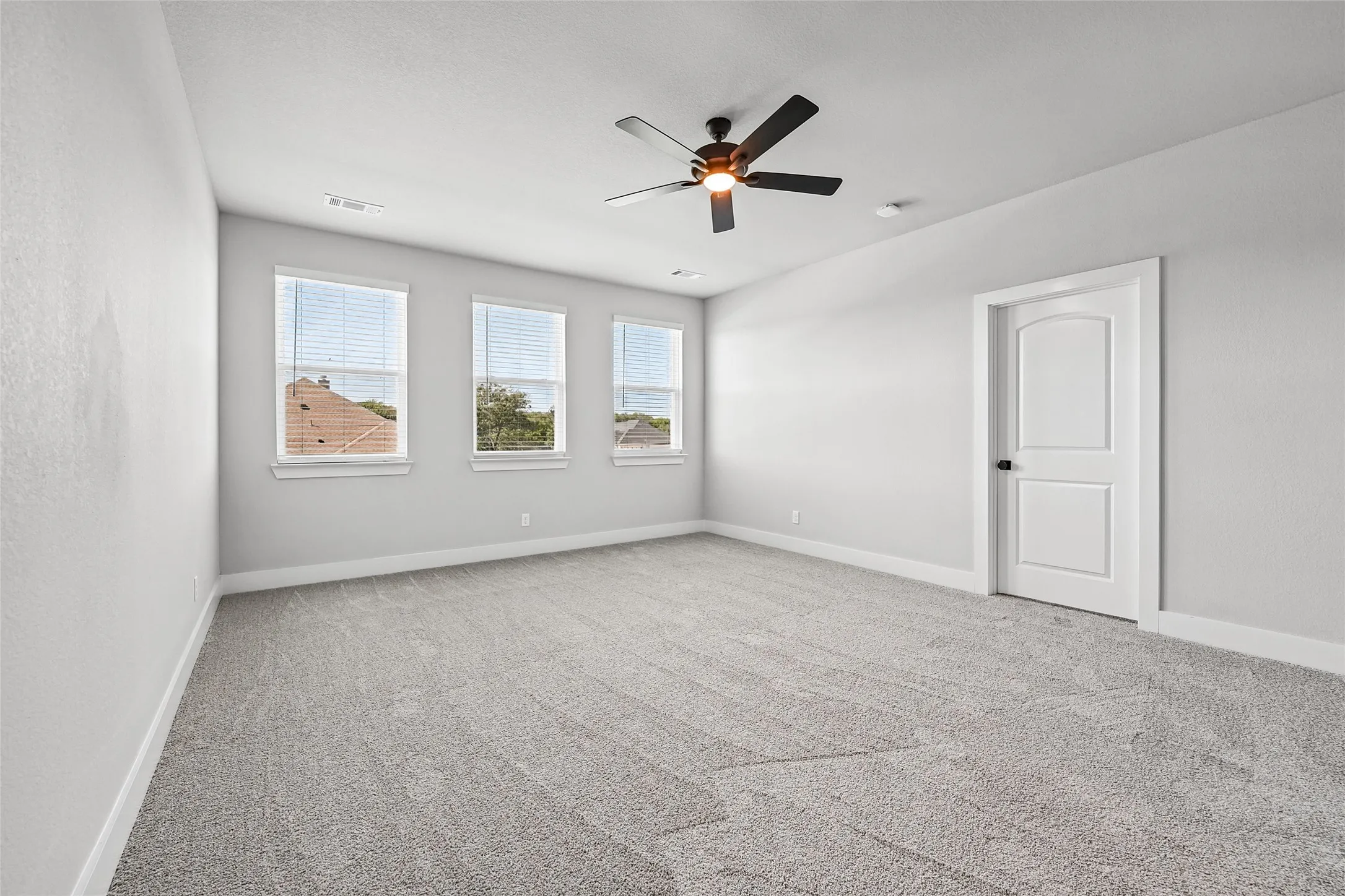 Neutral wall tones, three windows with blinds, ceiling fan with light fixture, light gray carpet, and a two-panel interior door