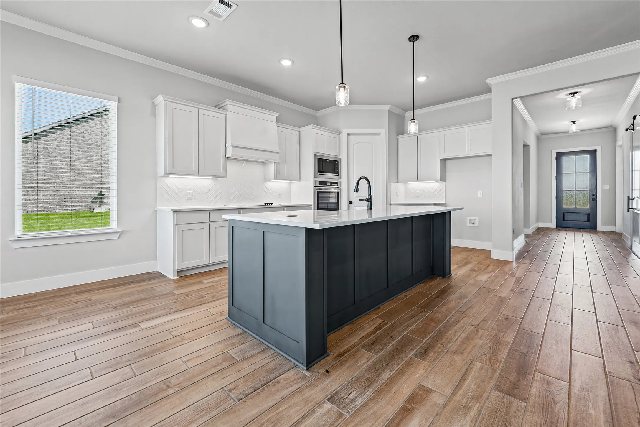 Wood-finish flooring throughout, white shaker cabinetry, dark kitchen island with stone countertop, built-in stainless steel appliances, and pendant lighting fixtures