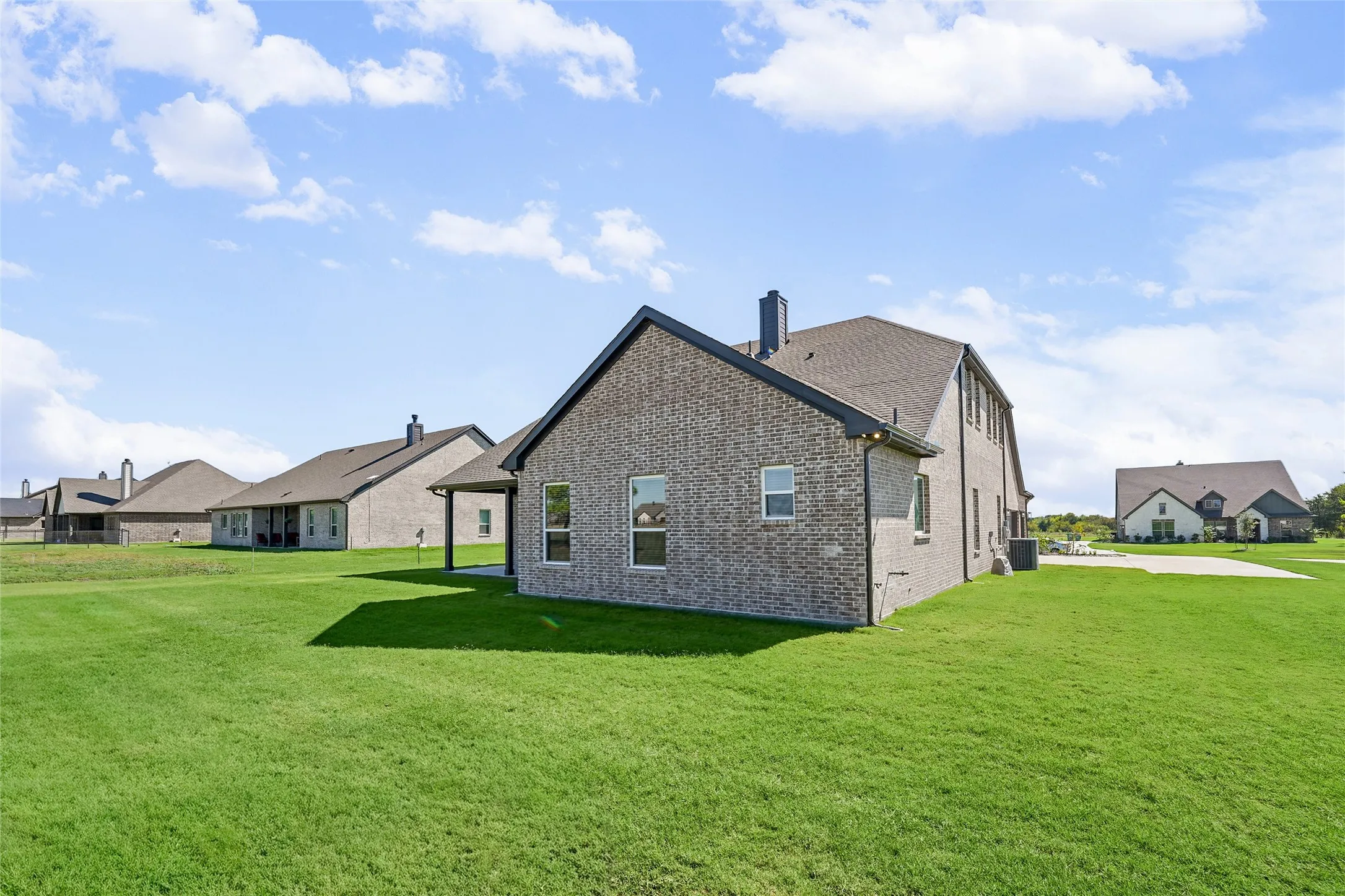 Expansive green lawn surrounding a brick exterior residence with a covered patio, multiple windows, and a dark shingled roof