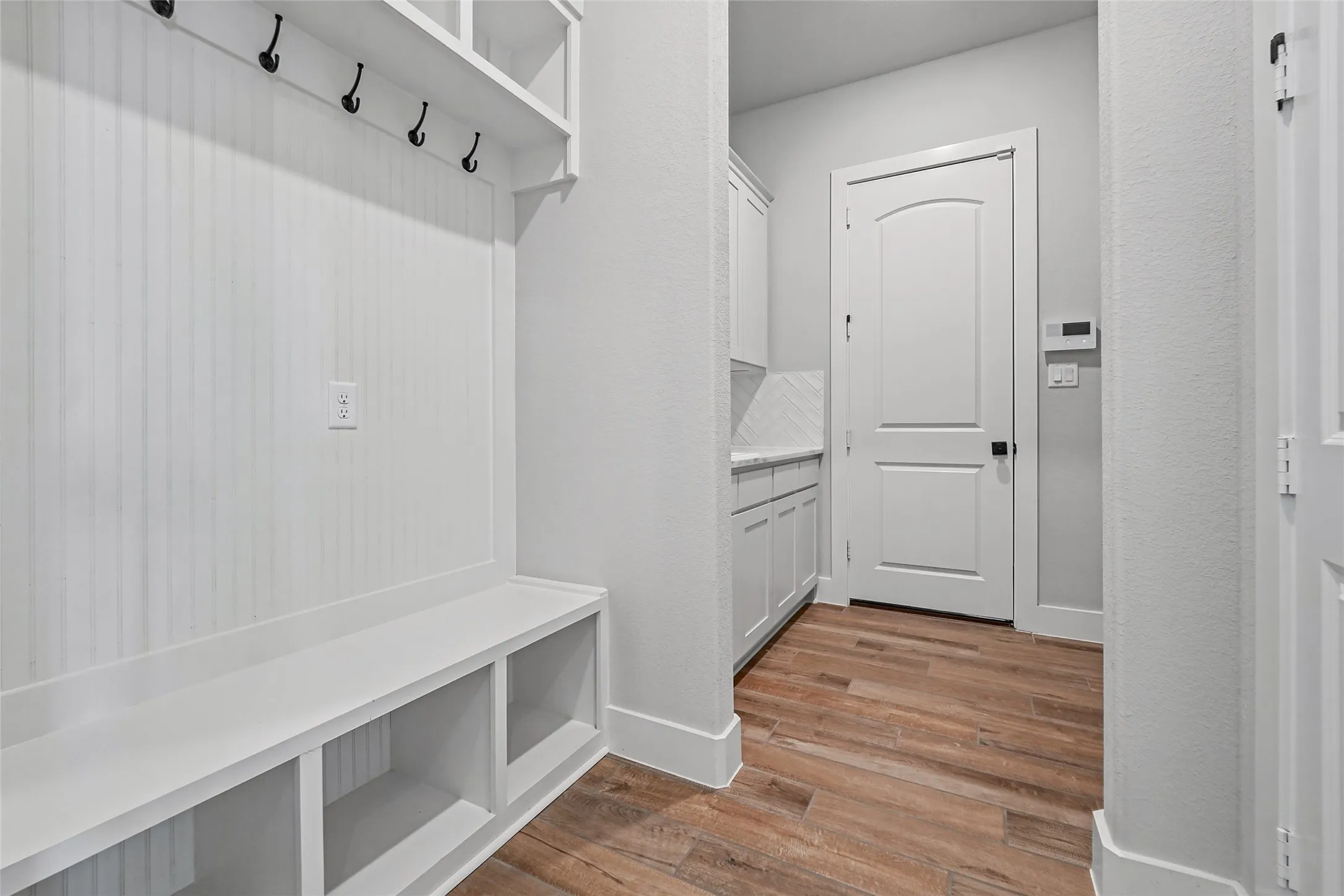 Built-in storage bench, overhead shelving with coat hooks, wood-finish flooring, white cabinetry with light-colored countertop, and a white interior door
