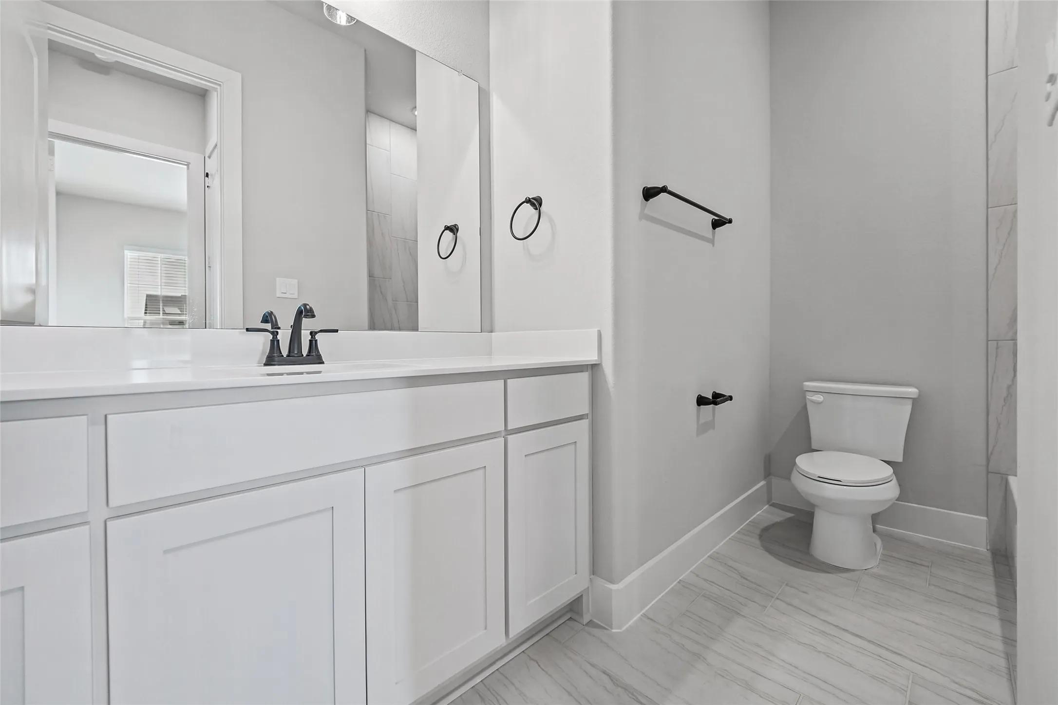 White vanity cabinetry with matte black hardware, white countertop, rectangular mirror, and light-toned tile flooring