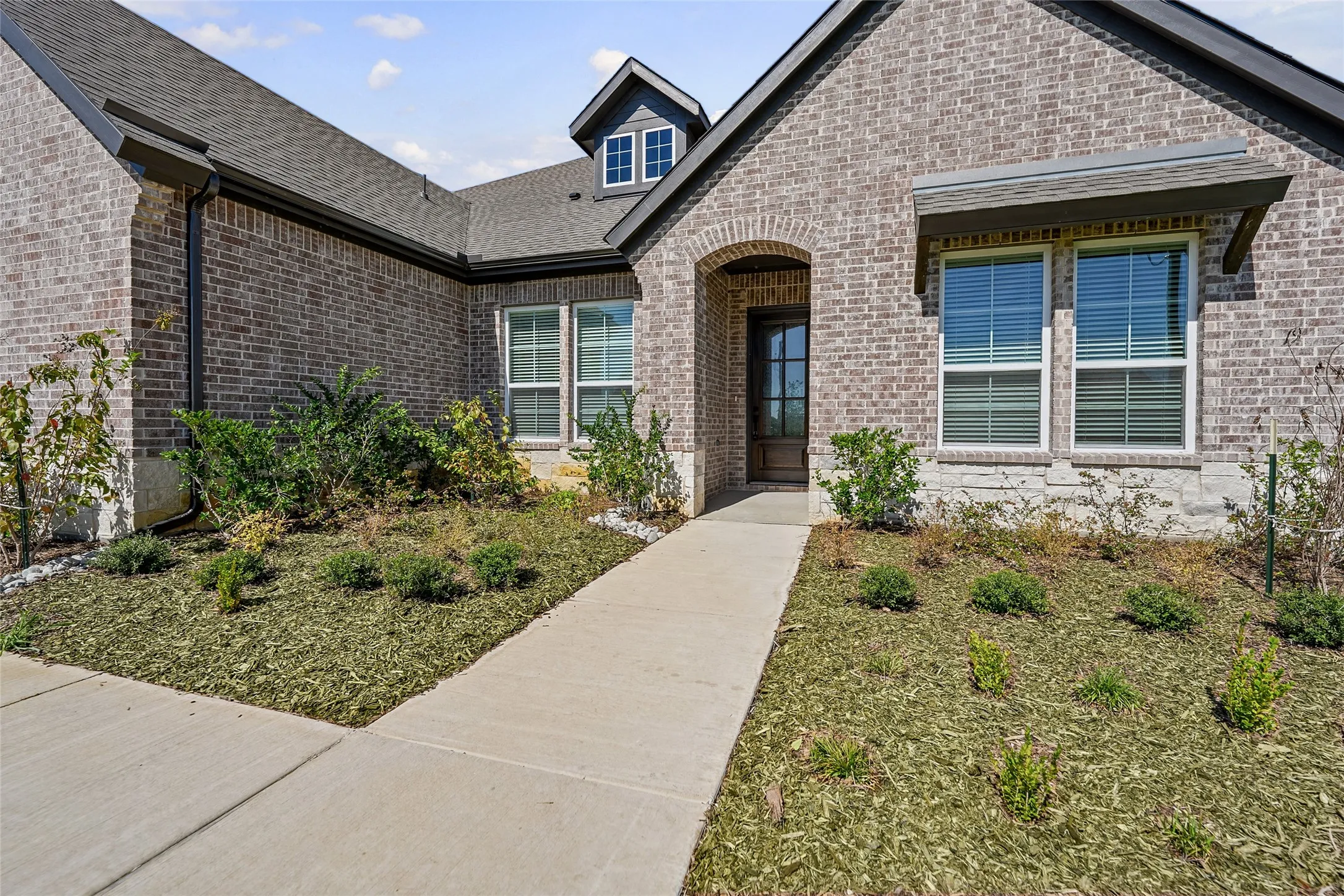 Brick and stone exterior, arched entryway, dark wood-finish front door, multiple windows with blinds, dormer window