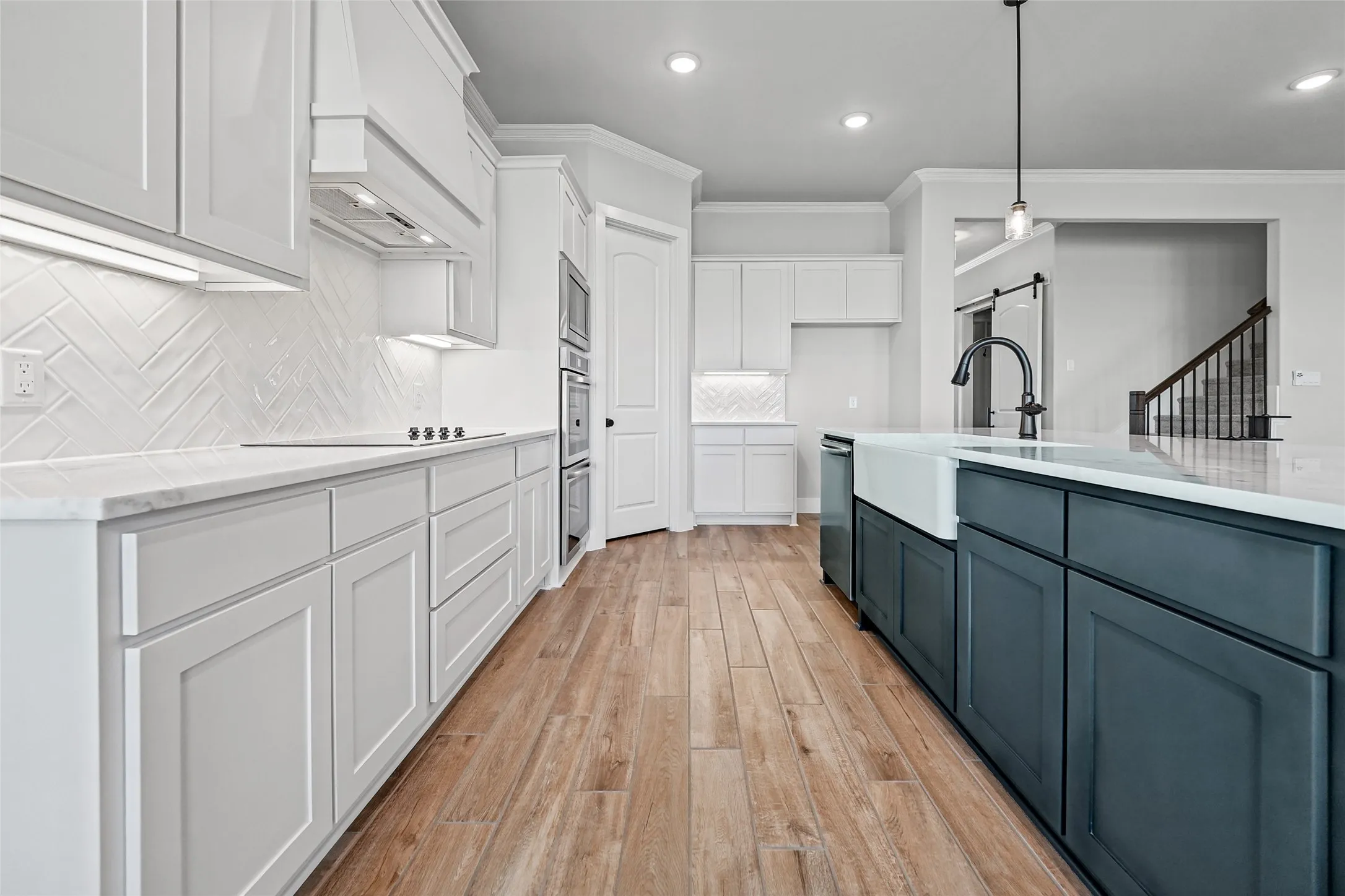 Kitchen featuring wood-finish flooring, white shaker-style cabinetry, a herringbone tile backsplash, a center island with a farmhouse sink, and stainless steel double ovens