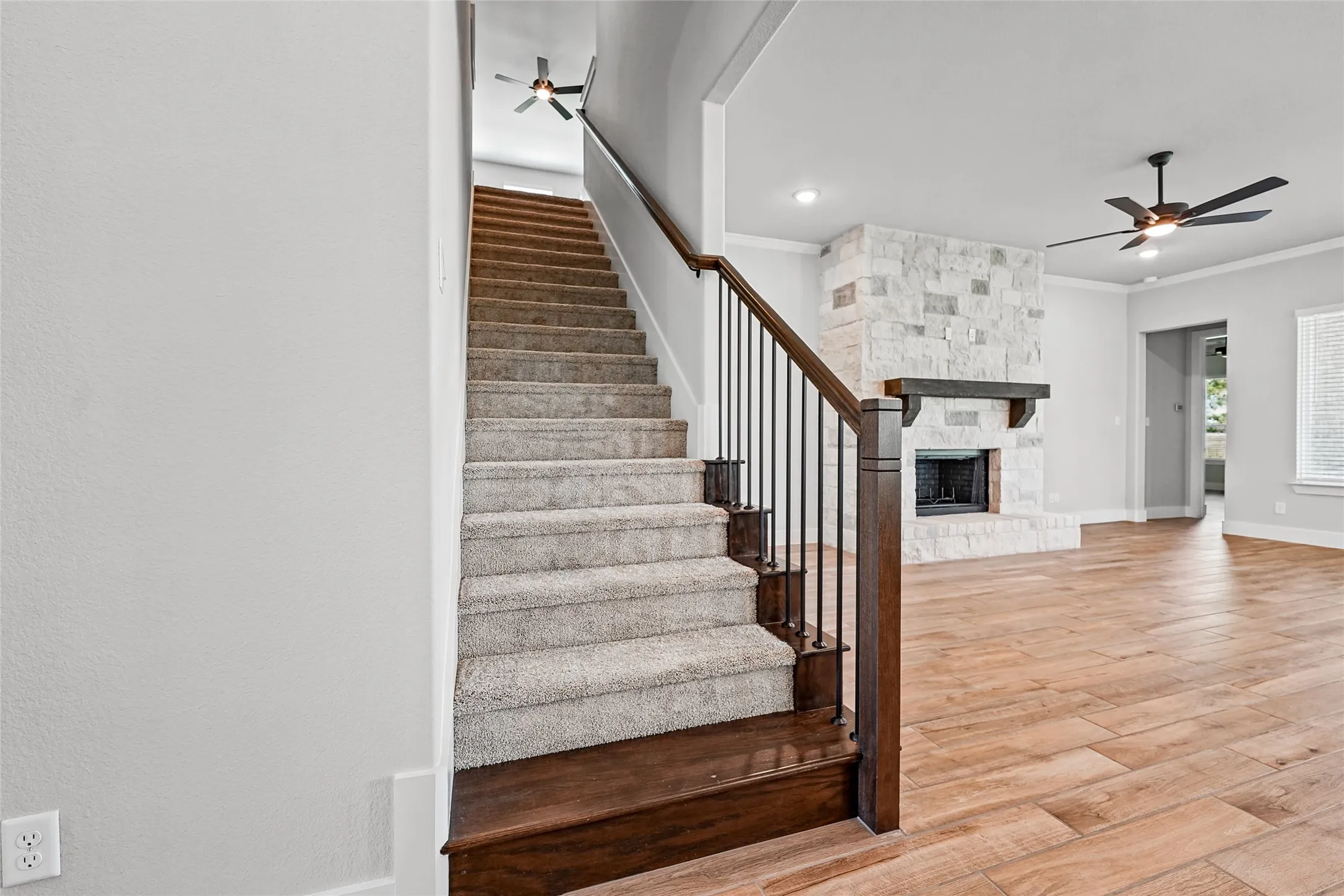 Wood-finish flooring, stone-clad fireplace with wood mantle, carpeted staircase with dark wood newel posts and metal balusters, ceiling fans