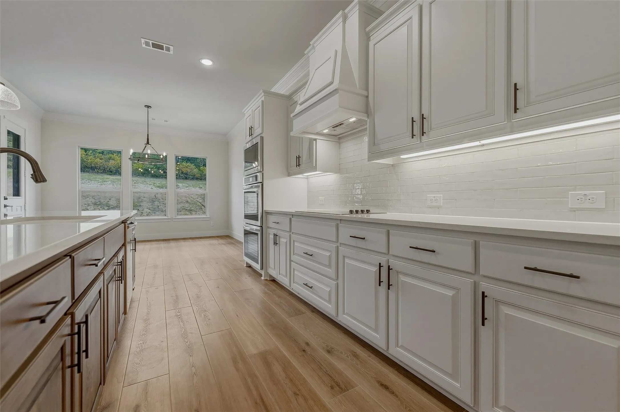 White shaker-style cabinetry with dark hardware, integrated stainless steel double oven, tile backsplash, and light wood-finish flooring