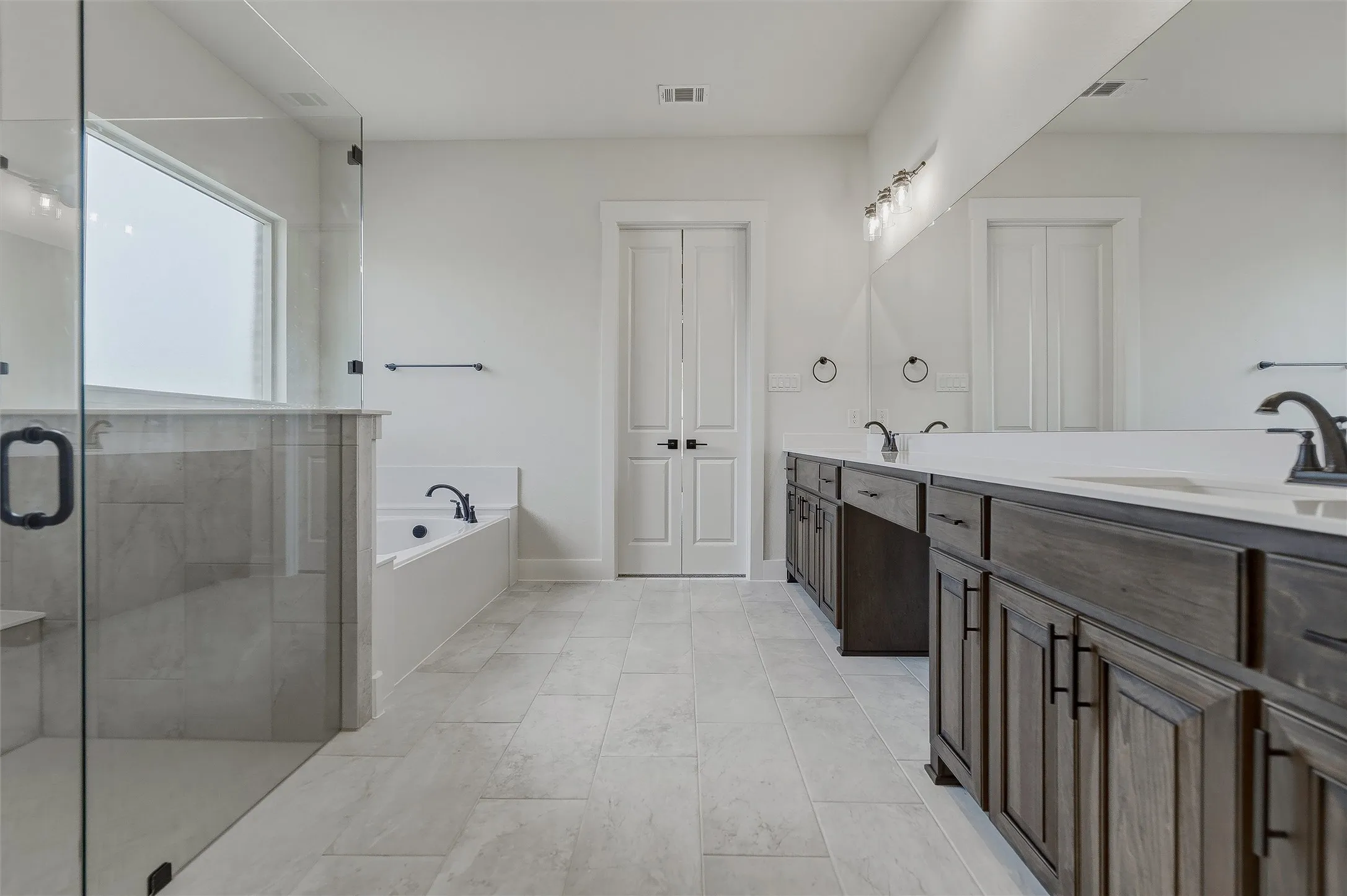 Spacious bathroom with glass-enclosed shower, built-in soaking tub, dual vanities with dark wood cabinetry, light countertops, and large format tile flooring