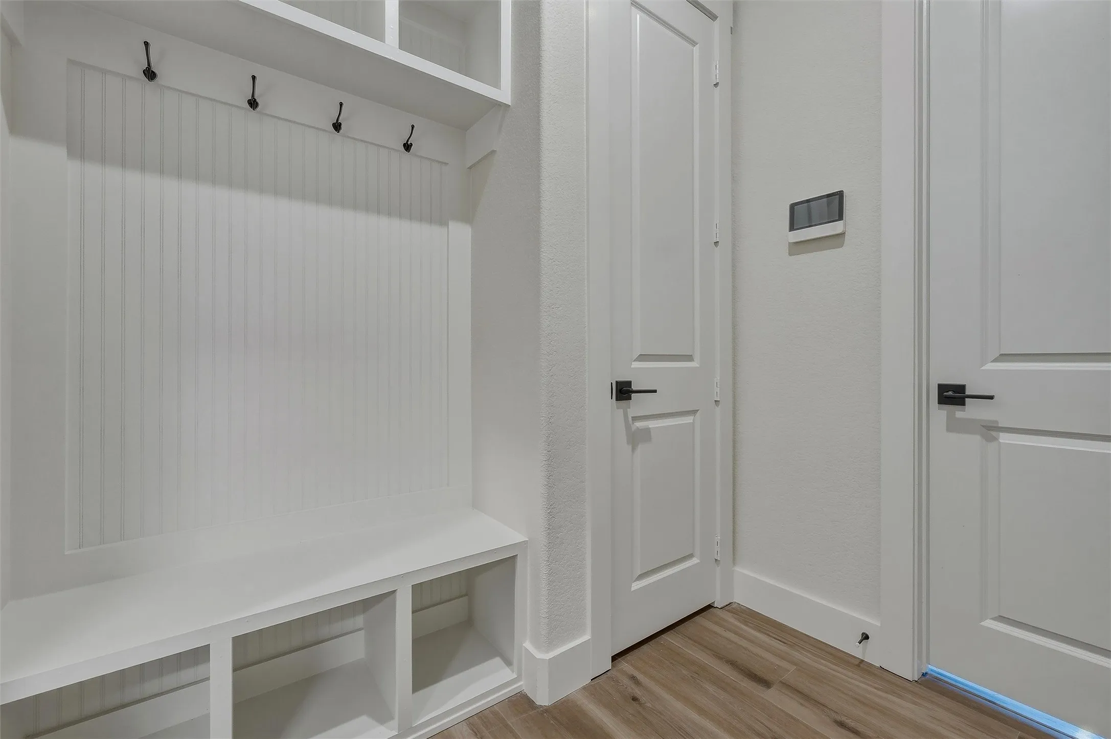 Built-in storage bench with cubbies, upper shelving, and beadboard paneling with hooks, complemented by wood-finish flooring and solid core doors with black hardware
