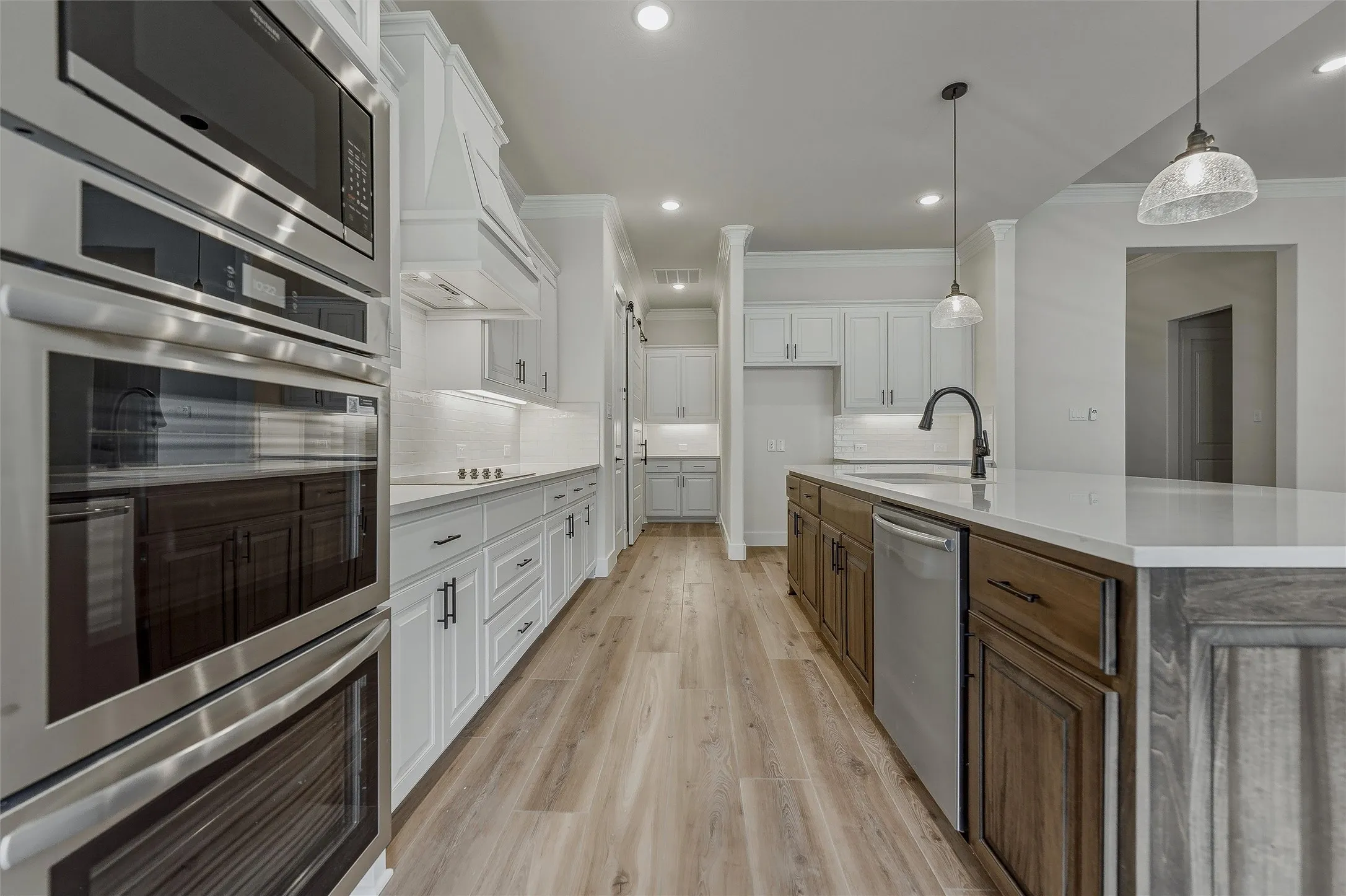 Kitchen featuring wood-finish flooring, white cabinetry, a built-in double oven and microwave, a center island with sink, and pendant lighting