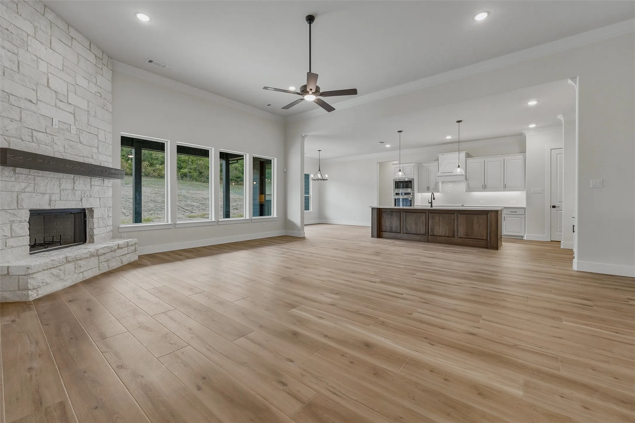 Stone fireplace with a wood mantel, wood-finish flooring, recessed lighting, and a kitchen island with pendant lighting
