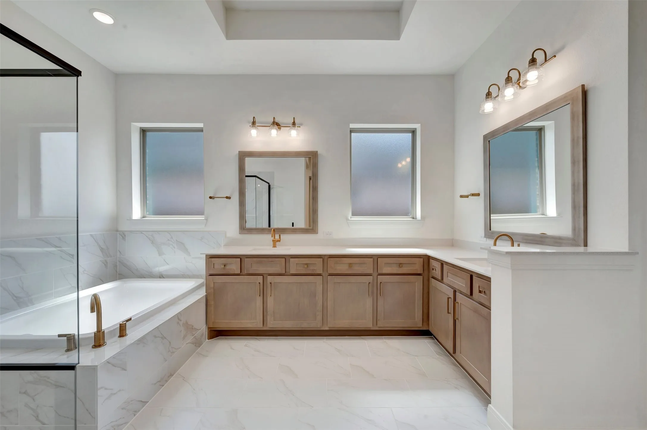 Spa-inspired primary bathroom featuring a wood-finish vanity with dual sinks, light stone countertops, and abundant natural light from multiple windows