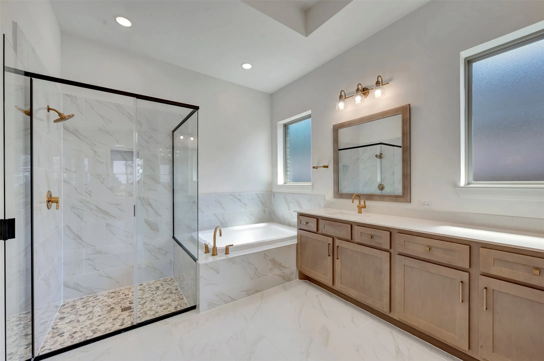 Spa-inspired primary bathroom showcasing a marble-clad walk-in shower with frameless glass enclosure and rainfall showerhead, freestanding soaking tub, dual-sink vanity with wood cabinetry and warm brass fixtures, and abundant natural light from multiple windows