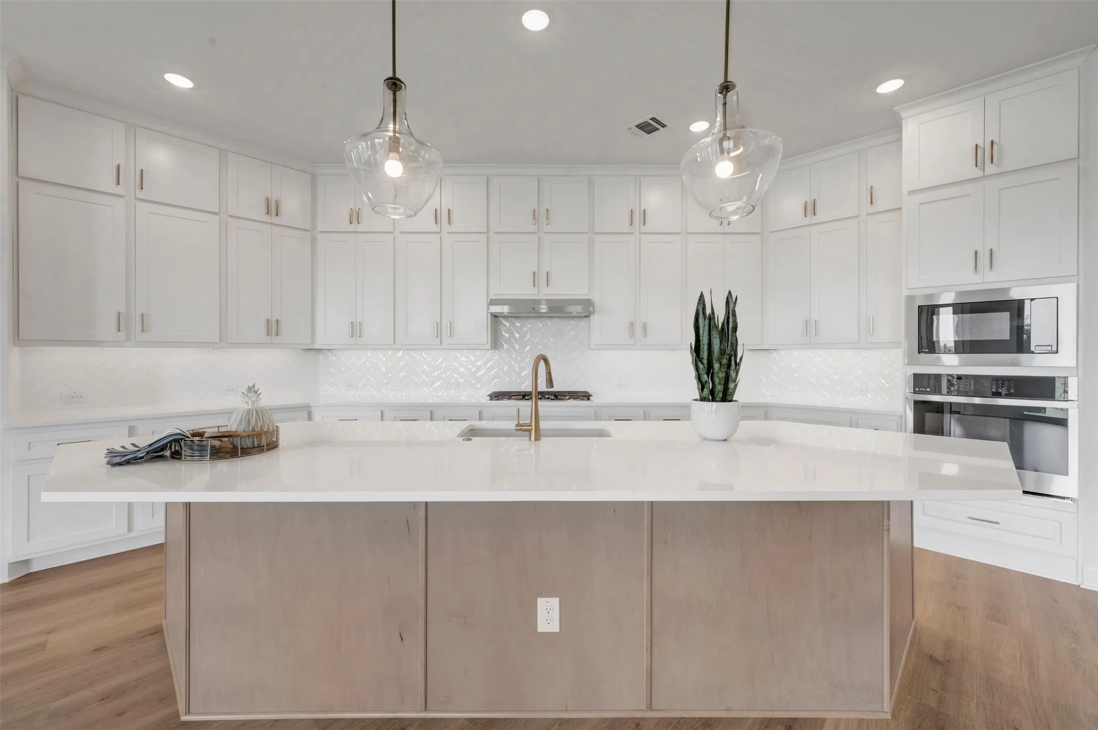 Chef's kitchen featuring white shaker cabinetry, a substantial wood-finish island with multiple storage solutions, quartz countertops, stainless steel appliances including a double wall oven, pendant lighting with glass shades, and a marble subway tile backsplash with integrated sink and bronze faucet