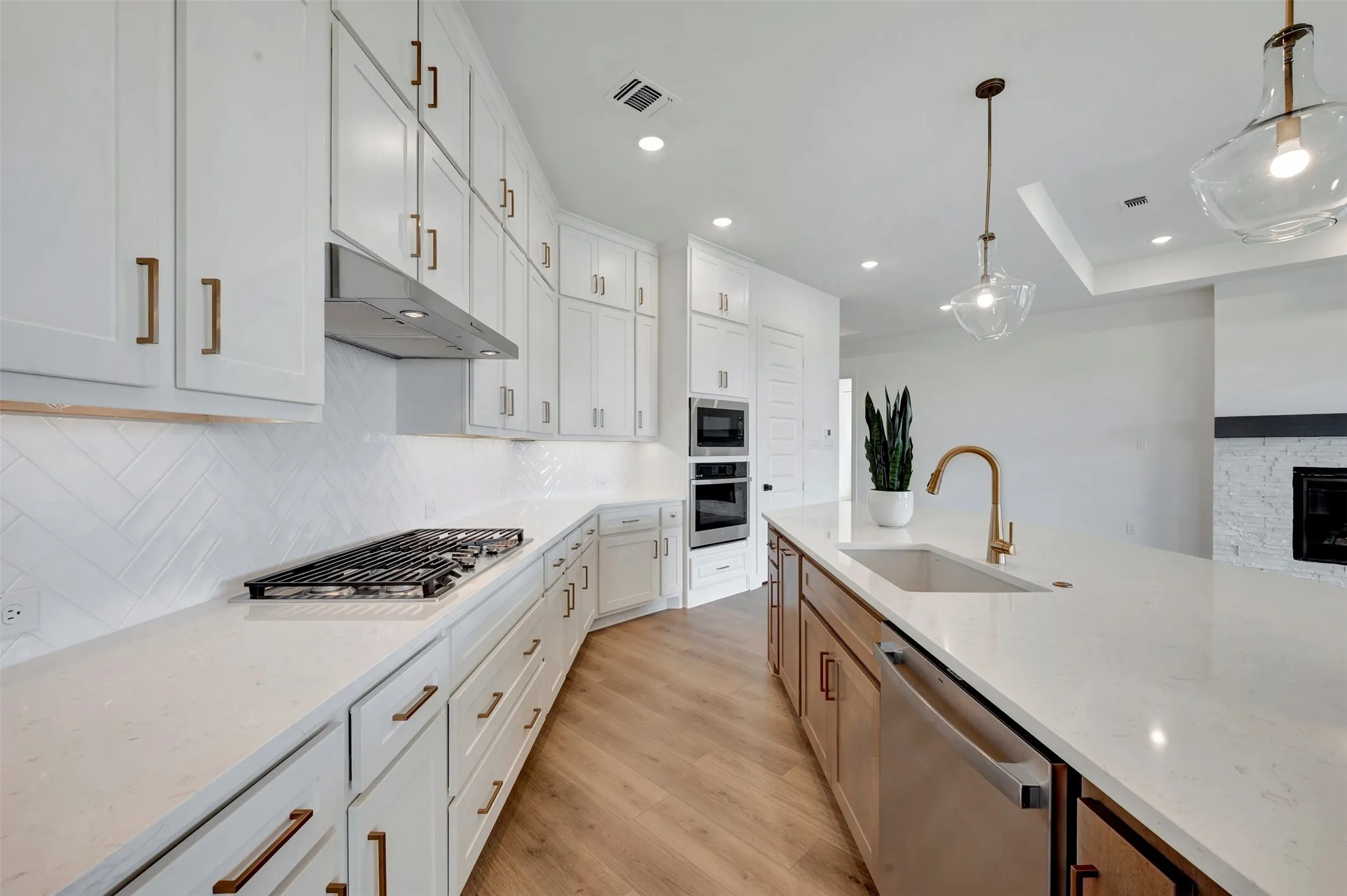 Modern kitchen featuring white cabinetry with brass hardware, white subway tile backsplash, stainless steel gas cooktop, dual wall-mounted ovens, quartz countertops, an oversized island with waterfall edge, brass gooseneck faucet, recessed and pendant lighting, and wood-finish flooring throughout
