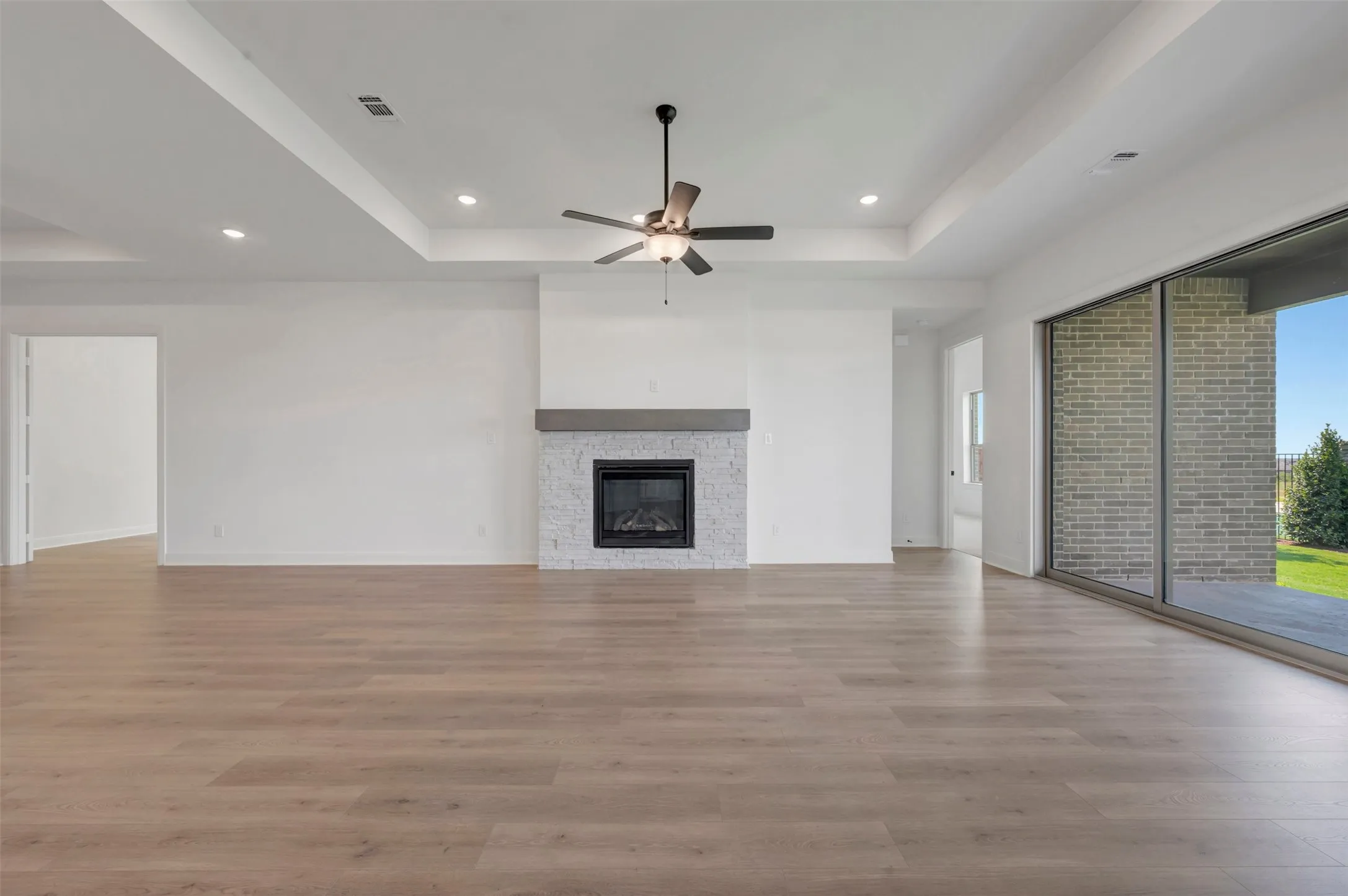 Spacious open-concept living area showcasing wood-finish flooring throughout, a coffered ceiling with recessed lighting and ceiling fan, a custom fireplace with gray tile surround, and expansive glass sliding doors opening to outdoor space