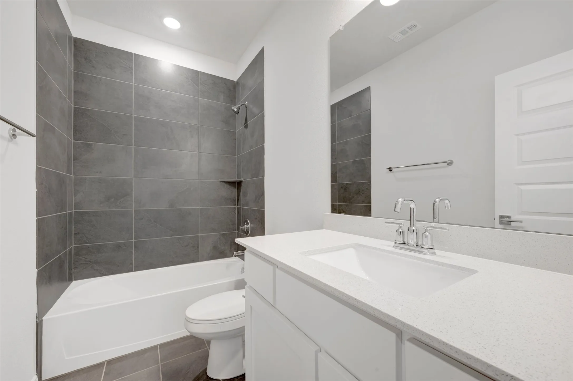 Bathroom featuring a white vanity with an integrated sink and light-colored countertop, chrome fixtures, a full-width mirror, and a white bathtub with dark gray tile surround