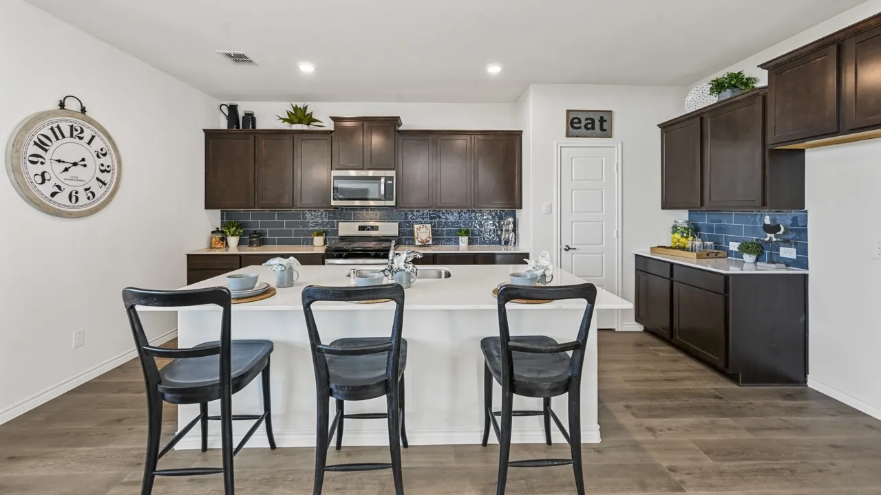 Spacious kitchen featuring an expansive central island with seating, wood-finish flooring, dark wood cabinetry, stainless steel appliances, and a blue subway tile backsplash