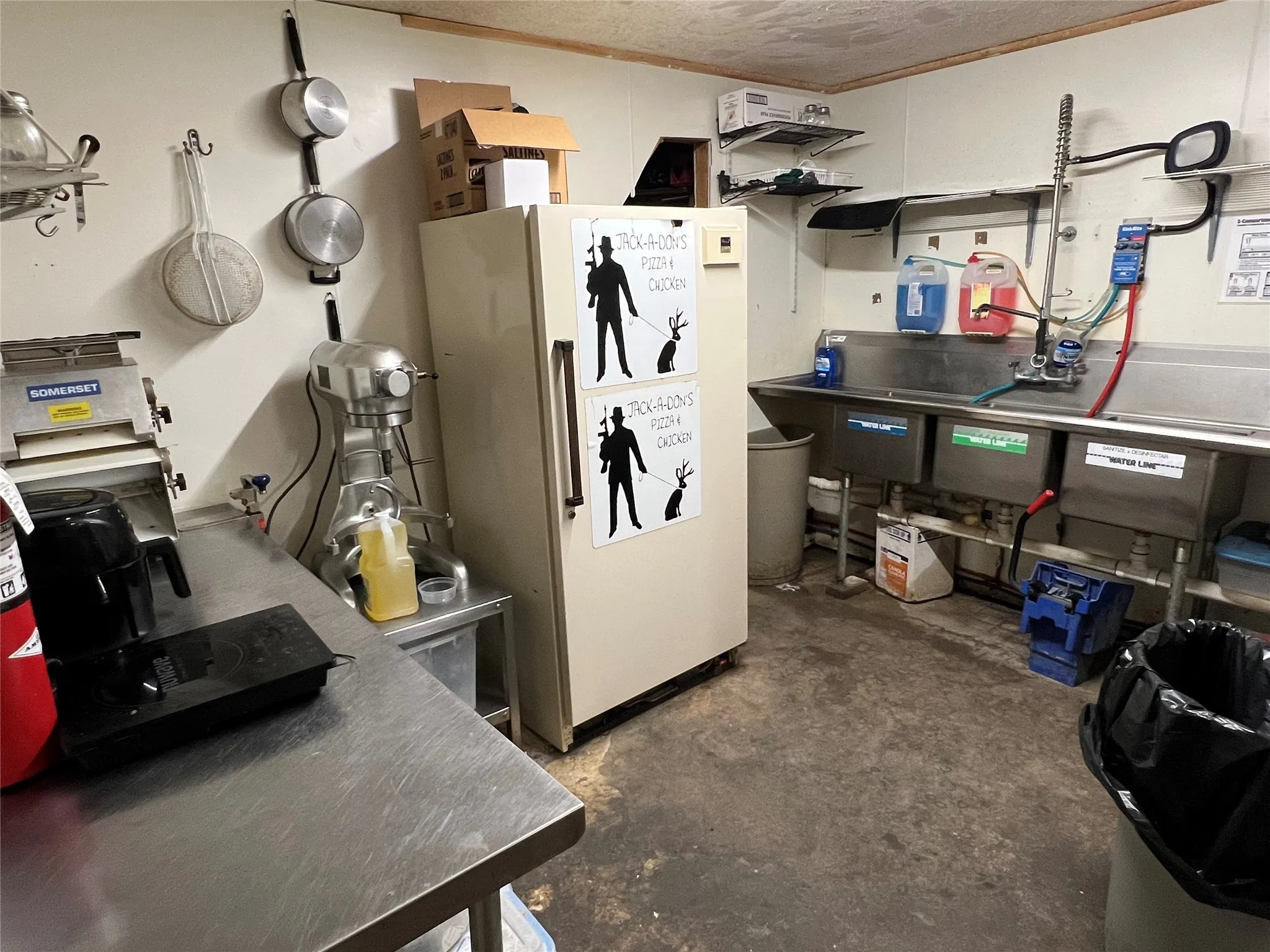 Commercial kitchen space featuring a triple-basin stainless steel sink, a commercial refrigerator, and stainless steel countertops